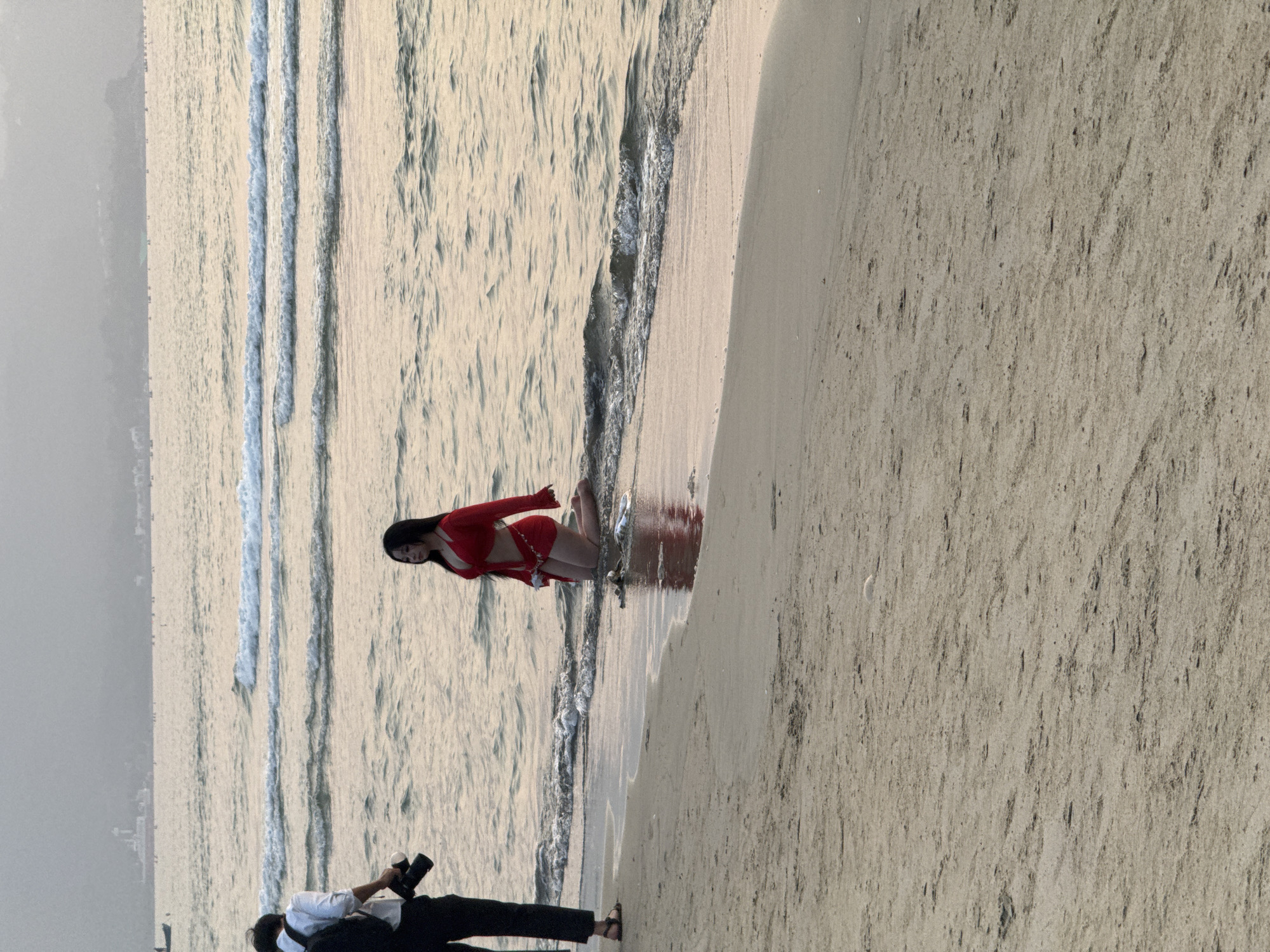 A coastal scene shows a woman in a red outfit posing at the beach's edge, with the ocean on one side and a sandy area on the other. A person with a camera stands on the sand, likely photographing the woman. The background includes ocean waves and a distant cityscape. The location is likely Đà Nẵng, Vietnam, based on the coastal scenery.