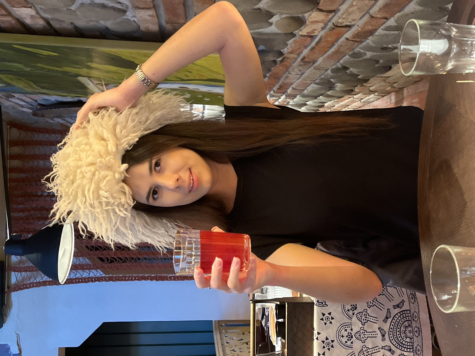 A young woman with long, dark hair and a fluffy white hat is sitting indoors. She is wearing a black t-shirt and a silver watch. She is holding a glass of red liquid in her right hand. The background consists of a brick wall and a painting of a landscape. There is also a wooden table and two glasses visible. The lighting suggests it is daytime, and the overall mood is casual and relaxed. The woman is smiling, and her eyes are looking towards the camera. The image is taken from a slightly high angle, looking down at the woman. The focus is on the woman and the glass of drink she is holding.