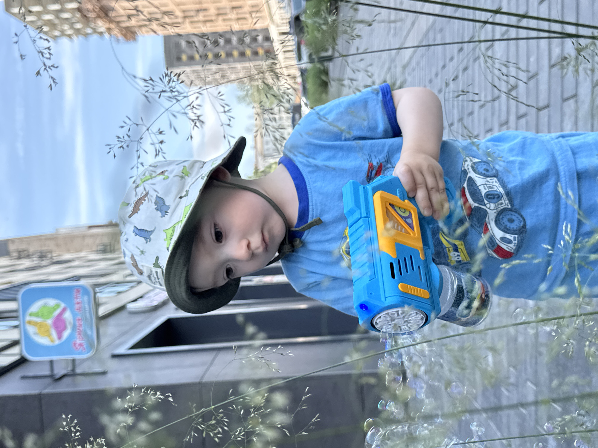In Izhevsk, Russia, a young Caucasian boy wearing a blue t-shirt with a car print and a bucket hat is playing with a bubble gun. He is standing on a paved sidewalk next to a building. Tall grass is in the foreground, obscuring some of the view. The sky is partly cloudy. Buildings and parked cars are visible in the background.