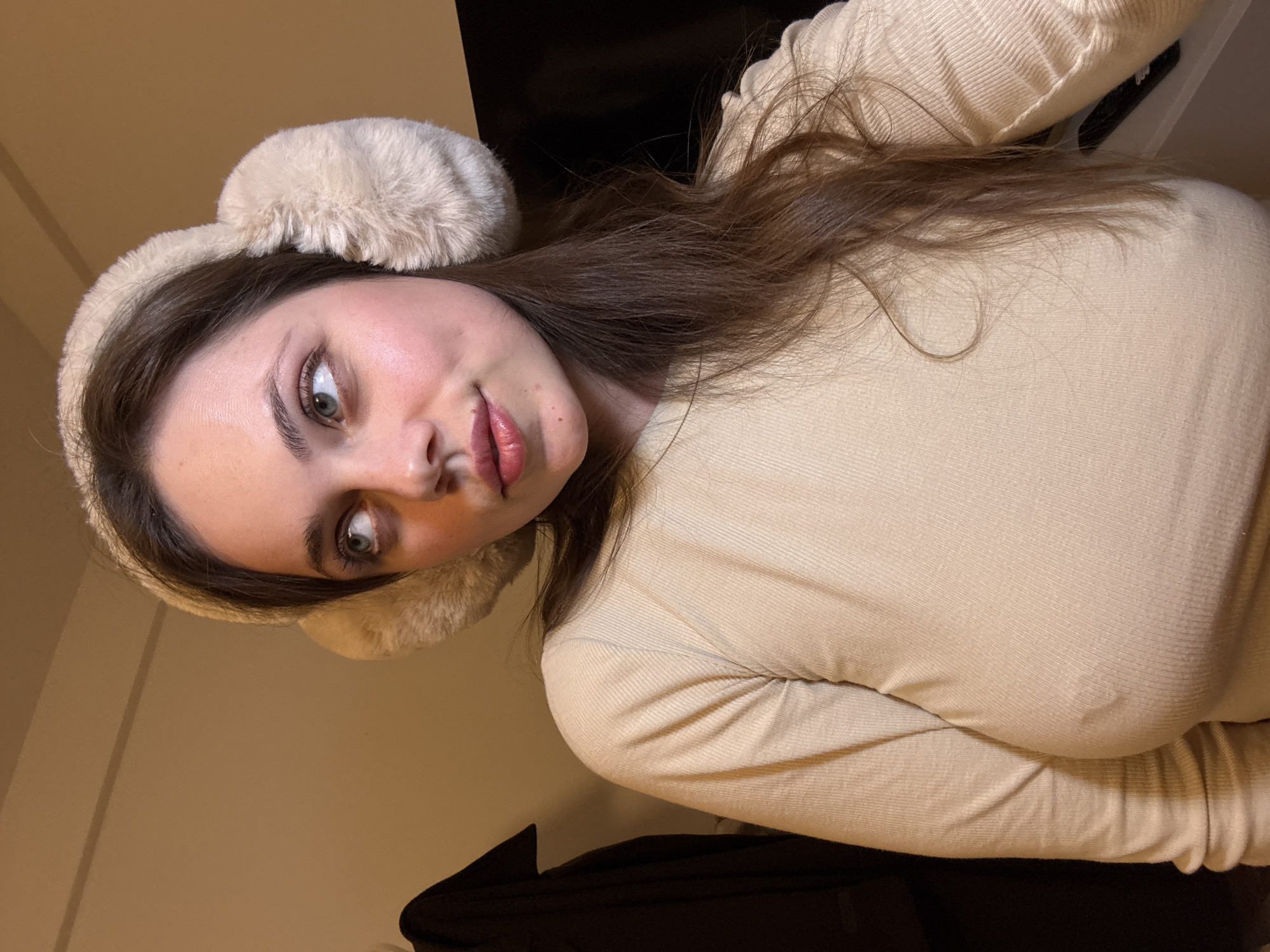 A young woman is shown in a close-up, angled self-portrait. She is wearing a cream-colored, long-sleeved top and a fluffy headband with ear-like protrusions. Her hair is brown and falls around her face. She has fair skin, light-colored eyes, and slightly parted lips. The background appears to be an interior space with a cream-colored wall and a dark object, possibly clothing or furniture, in the lower frame. There is no information in the image to confirm or deny that it was taken in Sappada / Plodn / Sapade, Italy.