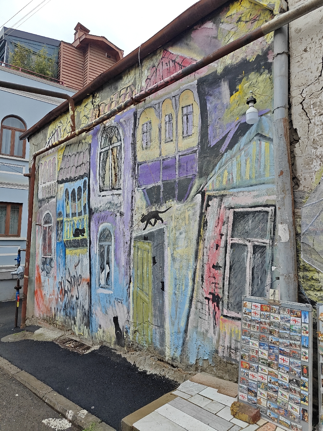The image presents a vibrant street scene, characteristic of Tbilisi, Georgia, centered around a large, colorful mural painted on the side of an old building. The mural depicts a whimsical row of houses rendered in bold, contrasting colors such as purple, yellow, blue, red, and light green, complete with windows and doors. Several black cat silhouettes are integrated into the artwork: one is shown walking along a roofline, another peeks from an arched window, a third sits near a yellow door, and several smaller cat shapes are scattered near the doorway. Two rusty horizontal pipes run across the upper part of the mural, and a vertical grey drainpipe is visible on the right, from which a small white light fixture hangs.
In the foreground to the right, a metal display stand is filled with numerous souvenir magnets or postcards, many prominently featuring the red and white flag of Georgia, clearly indicating the location. Below the stand, there are light-colored paving stones and a loose brick. A section of freshly laid black asphalt occupies the lower left of the frame, marking a path or road.
To the left of the mural, part of a blue-grey painted building with arched, brown-framed windows is visible. Above it, glimpses of a modern building with dark paneling and green foliage, along with a distinctive small, wooden structure with a gabled roof, can be seen in the background. The wall bearing the mural shows signs of age with cracked plaster, providing a textural contrast to the smooth, brightly painted artwork.
The scene takes place during daytime under an overcast sky, suggested by the soft, diffused lighting and absence of strong shadows. There is no visible human activity, but the souvenir stand implies a pedestrian area. Some partially obscured graffiti text, possibly "Tbilis," is visible on the lower left of the mural. The overall atmosphere blends artistic creativity with historic urban fabric.