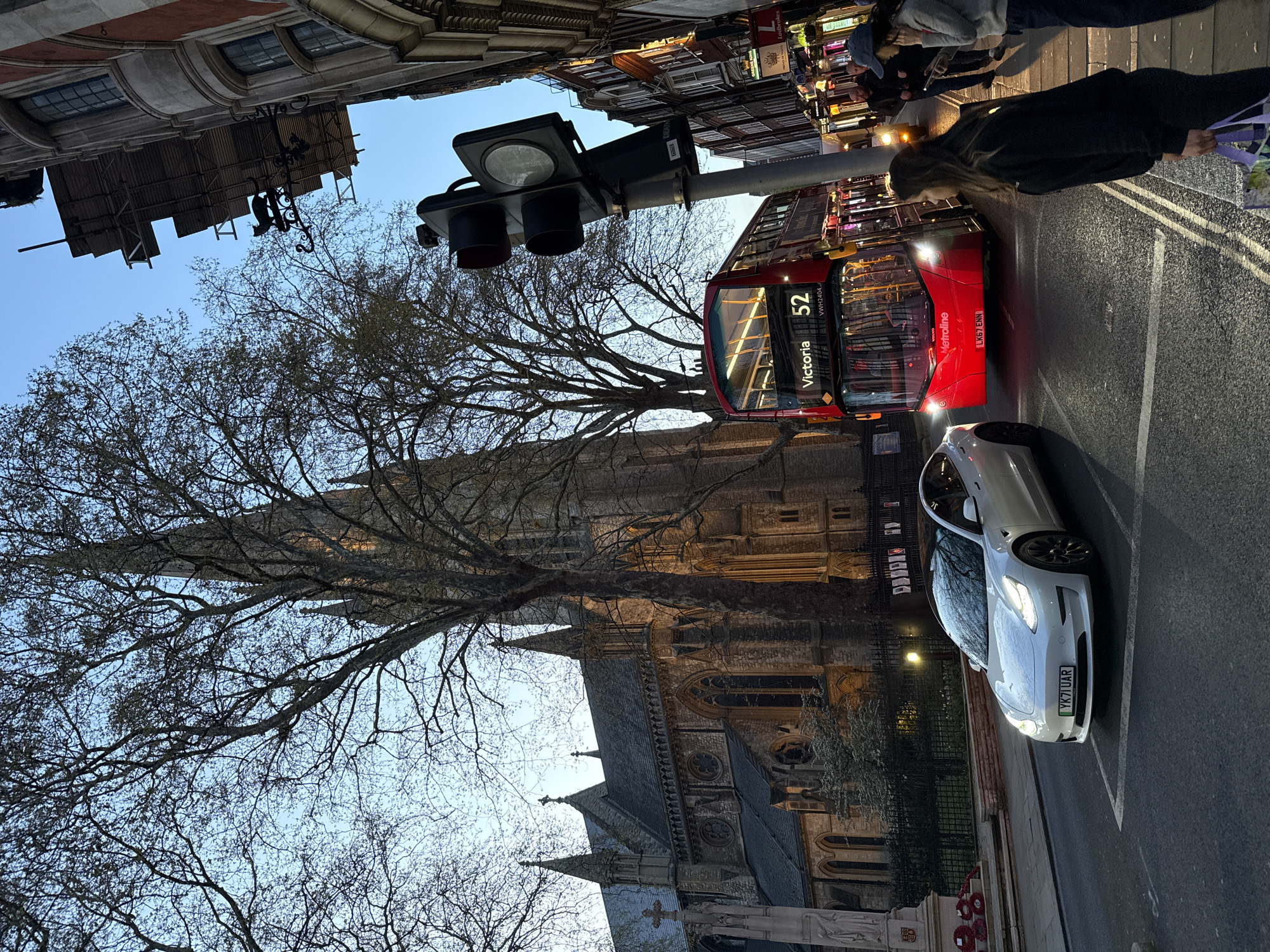 The image, taken from a low-angle perspective, shows a street scene in London, United Kingdom. In the foreground, the roof and spires of Westminster Abbey are visible behind bare trees. A red double-decker bus labeled "52 Victoria" is driving down the street, next to a white Tesla parked on the side of the road with license plate "YK71 UAR". A traffic light is suspended above the street. Pedestrians can be seen on the sidewalk in the upper right. Buildings line both sides of the street. The sky is a pale blue, suggesting it might be dawn or dusk.