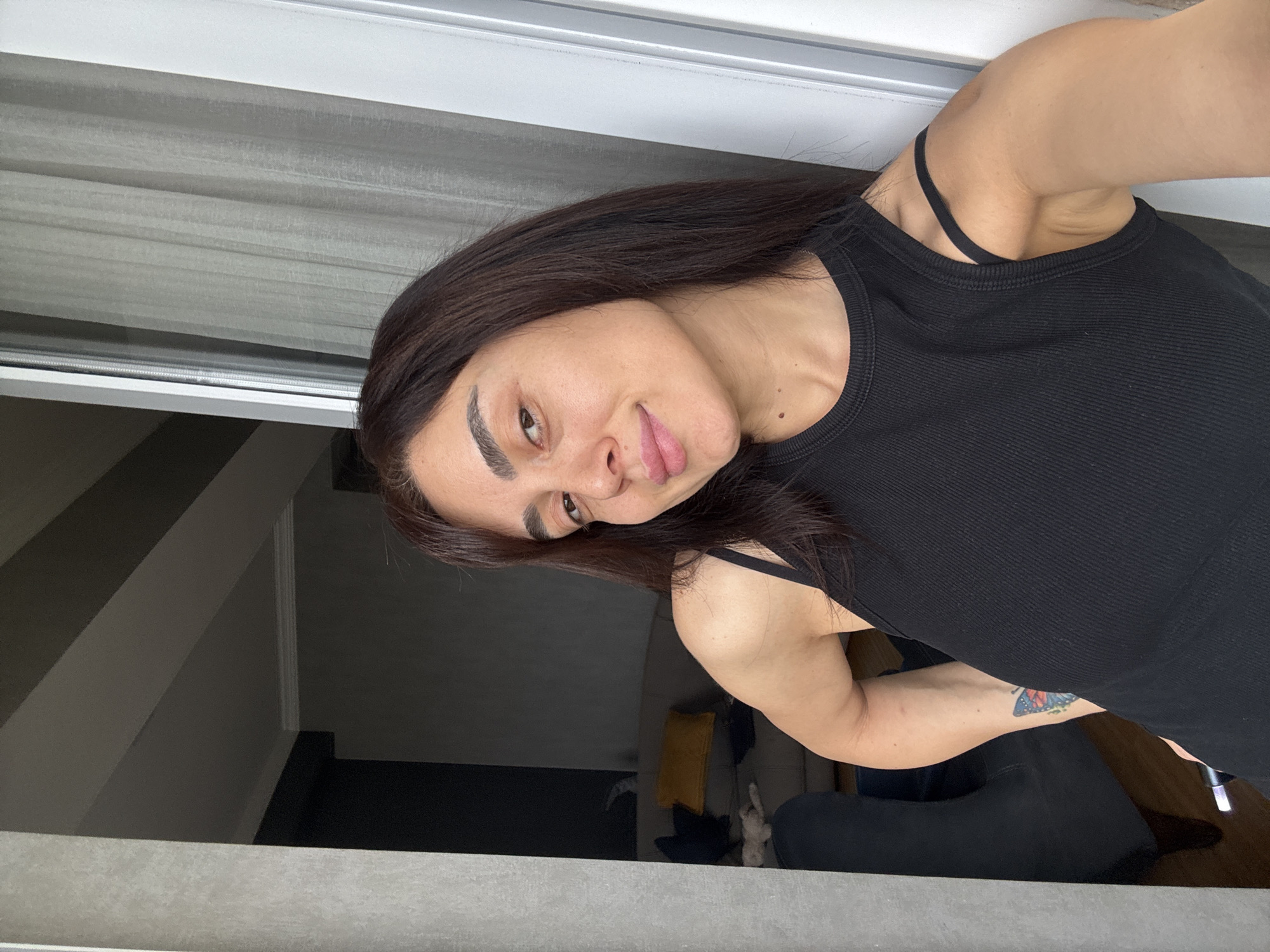 A person with dark hair is taking a selfie in what appears to be an indoor setting. They are wearing a black tank top. Their face is turned towards the camera, with their lips slightly parted and their head tilted. The background shows a grey wall and a window with blinds. Part of a staircase is visible in the background, suggesting the person might be on a higher floor or at the top of a staircase. A tattoo of a butterfly is visible on their arm. The lighting suggests it might be daytime. The overall mood appears casual. There is no visible text in the image.