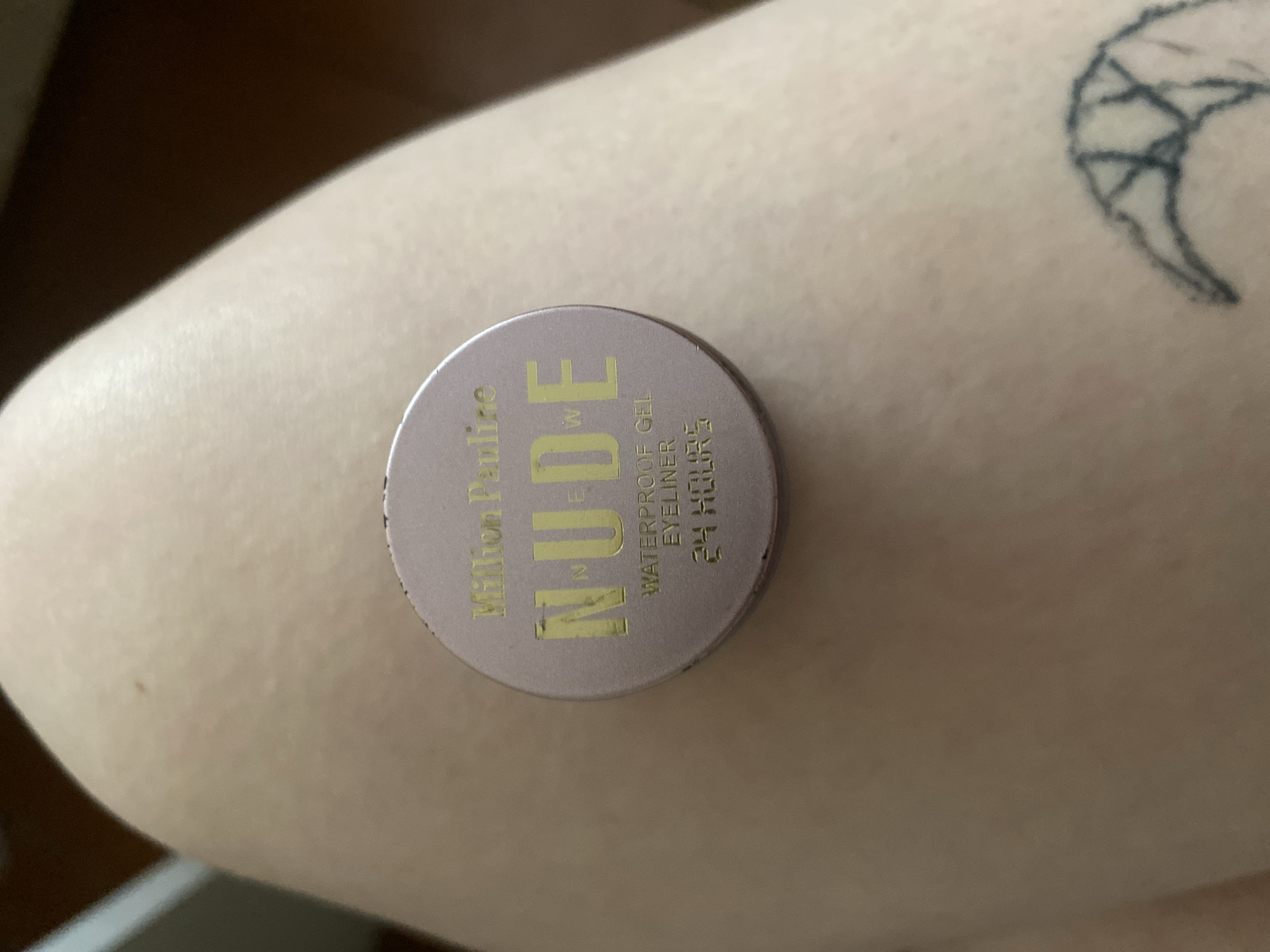 A small, round, light pink cosmetic container rests on a person's arm. The container has gold lettering that reads "Million Pauline NUDE WATERPROOF GEL EYELINER 24 HOUR." A tattoo of a crescent moon-like shape with a webbed interior is visible on the upper right part of the arm. The background is out of focus. There is no evidence in the image to suggest this is located in Chaikovsky, Russia.