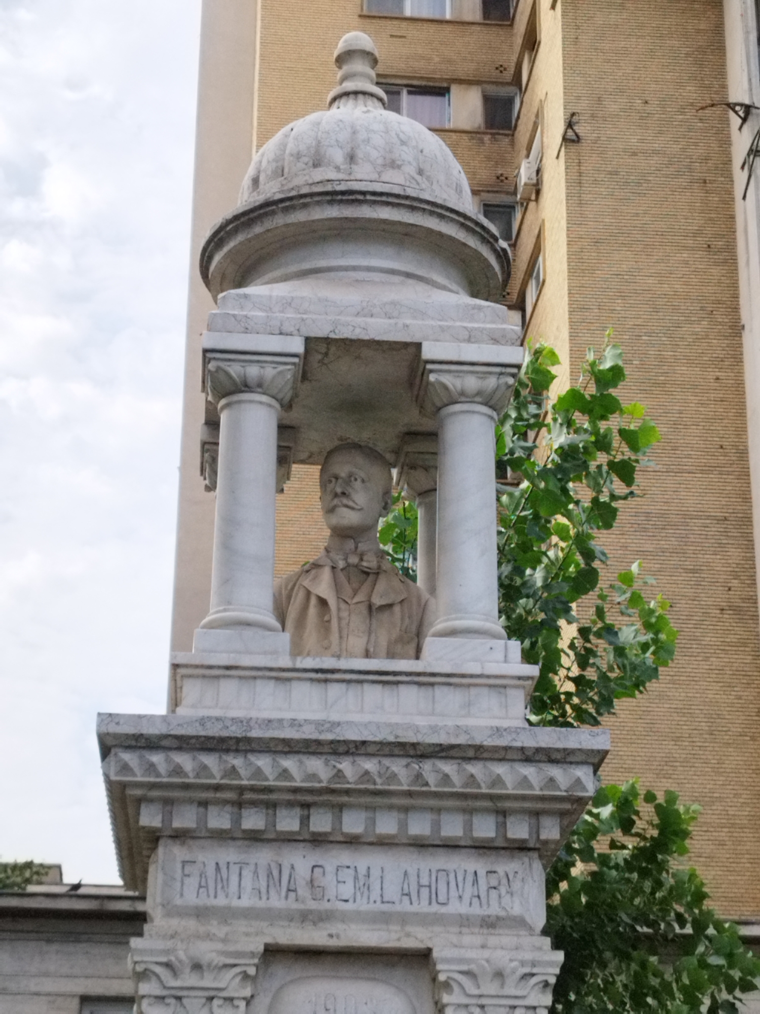 The image shows a marble bust of a man with a mustache and a bowtie, enclosed within a white marble monument. The monument features ornate carvings and pillars. The inscription "FONTANA G. EM. LAHOVARY" is visible on the monument. Behind the monument, a tall yellow brick building with windows is visible, along with lush green foliage. The sky is overcast. This monument is located in Bucharest, Romania, specifically in Cișmigiu Gardens, and it is dedicated to Grigore Em. Lahovary.