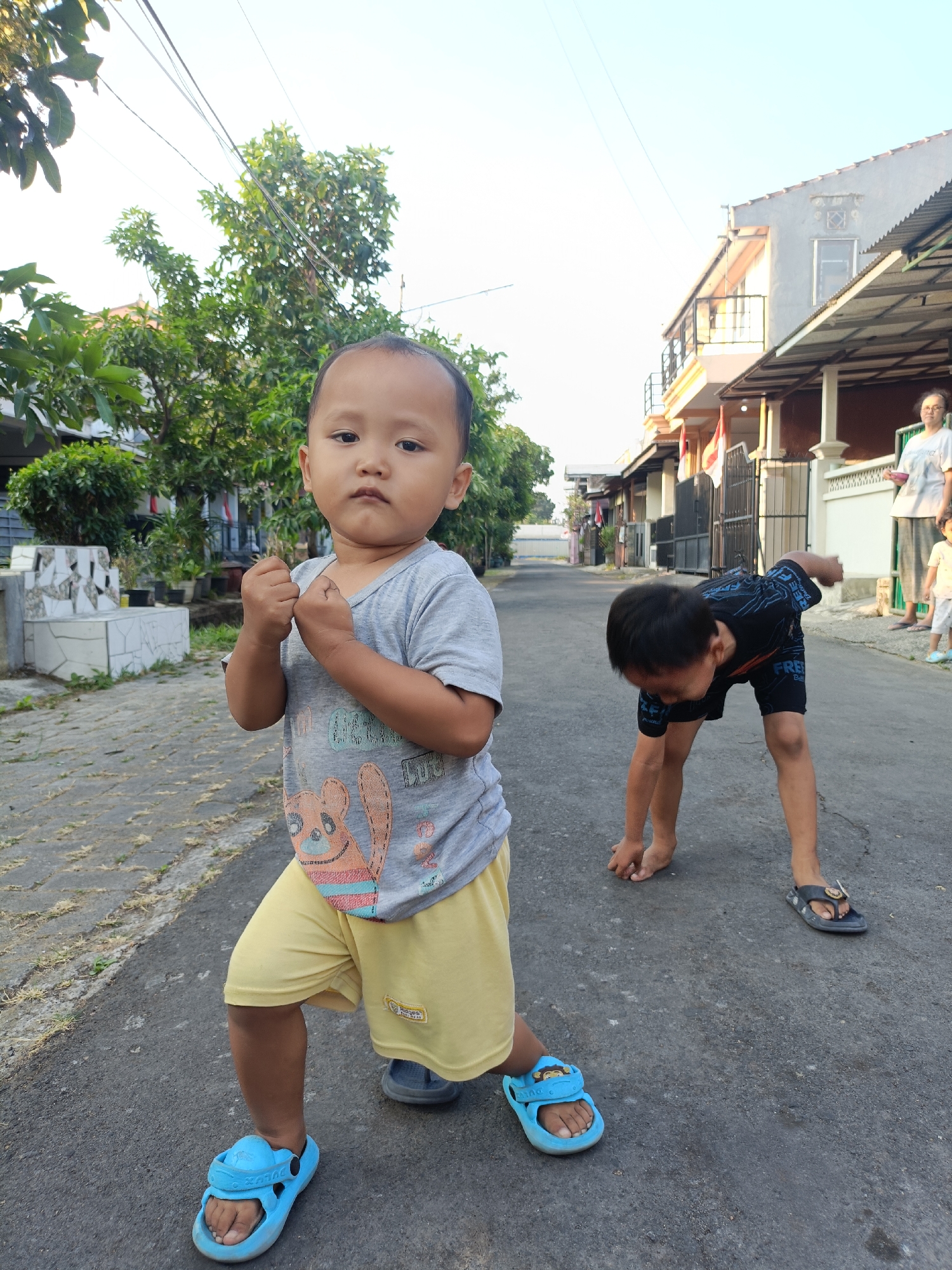 In a residential street in Kediri City, Indonesia, a young boy stands front and center, posing with his fists clenched. He wears a grey t-shirt with a cartoon bear graphic and yellow shorts, completed with bright blue sandals. To his right, another boy is bent over, seemingly engrossed in an activity on the asphalt road. In the background, houses line the street, with trees and vegetation adding to the urban landscape. A woman and a child are visible further down the street, near the houses. The sky above is clear and bright, indicating daytime.
