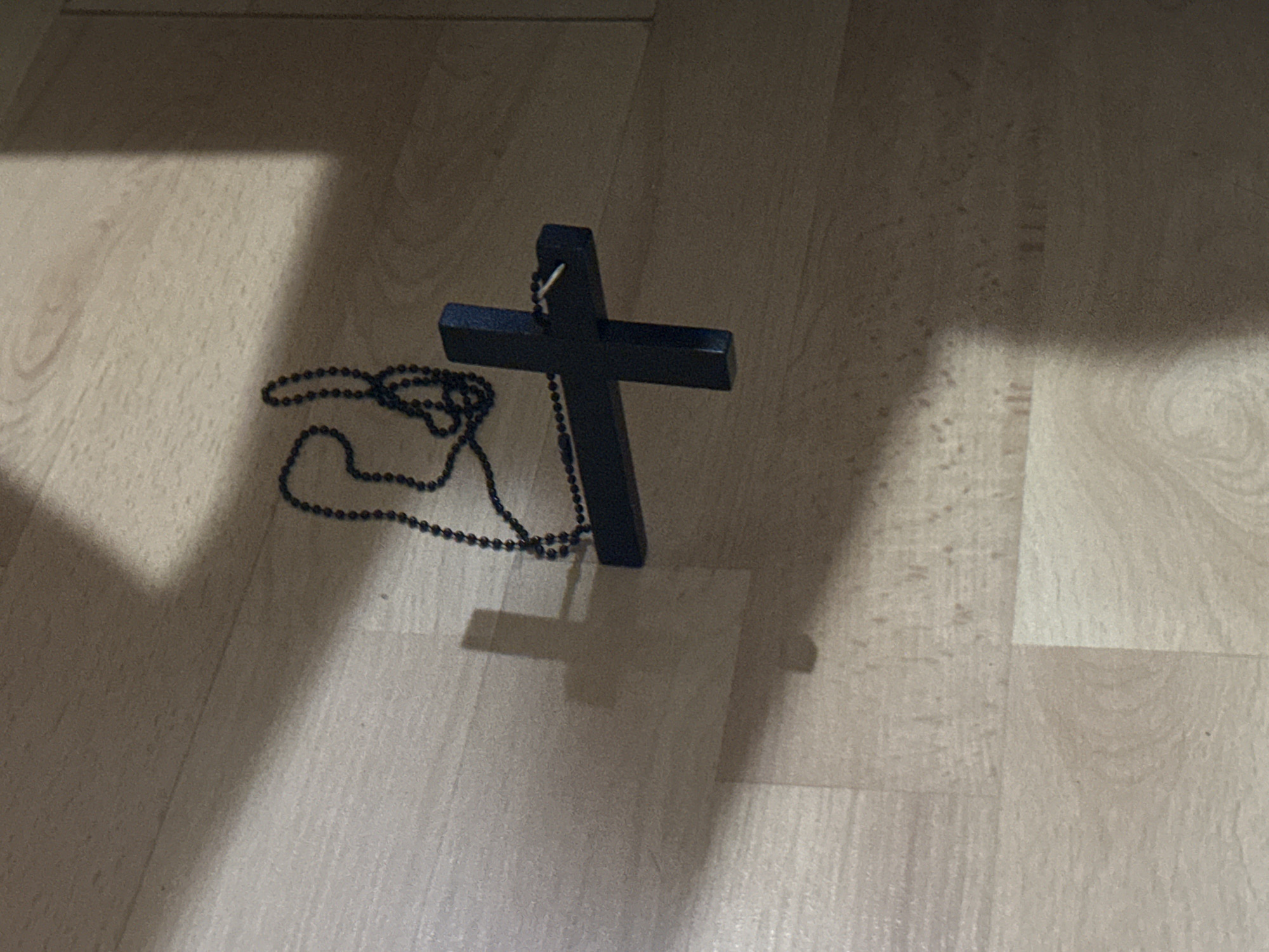 A black cross pendant with a beaded necklace is lying on a light-colored laminate floor. The image appears to be taken indoors, with light streaming in from the left, creating shadows. The floor has a wood grain pattern. There is no information in the image to identify the location as Moscow, Russia.