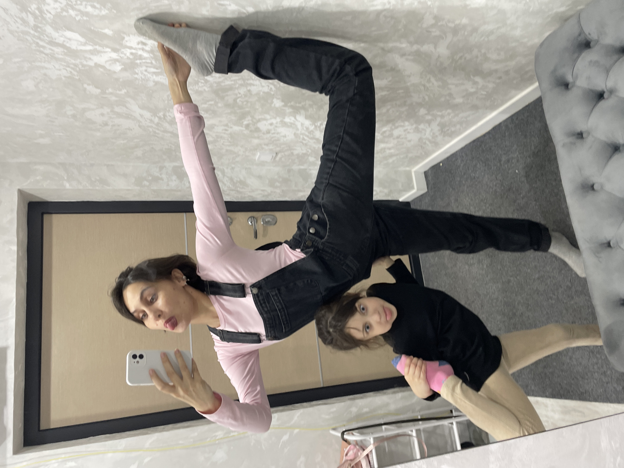 This media file captures a playful mirror selfie featuring an adult woman and a young child in an indoor setting. The image is taken as a reflection in a large mirror, showing the room and its occupants.
The adult woman, positioned on the left side of the frame (after conceptual rotation), has dark hair and wears a pink long-sleeved top underneath black overalls or dungarees. She is performing an acrobatic or stretching pose, standing with her left leg extended straight upwards, her grey-socked foot pressing against the ceiling. She holds a white smartphone in her left hand, capturing the reflection, and looks towards the camera with a playful expression, her tongue sticking out.
To the right, a young child, also with dark hair, is sitting or kneeling on the dark grey carpeted floor. The child wears a black long-sleeved top and light beige leggings, with a pink sock visible on one foot. They hold a small blue and pink object, possibly a toy or bottle, and look towards the camera with a neutral gaze.
The room features a light-colored wall or ceiling with a subtle wavy pattern. Behind the woman, a light brown wooden door with a prominent black frame is visible. On the far right, part of a grey tufted sofa or armchair is present. The overall setting suggests a modern home interior, possibly in Izhevsk, Russia. The time of day is not evident, but the scene is brightly lit indoors. There is no visible text.