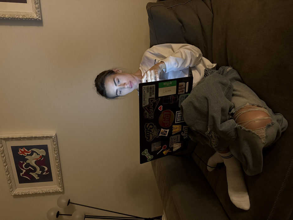 A young adult female is seated on a dark-colored upholstered couch in a dimly lit indoor residential setting, identified as a location in Venice, Italy. She is engaged with a dark-colored laptop, the screen of which emits a bright light that illuminates her face and hands. The laptop's lid is extensively covered with various stickers, including recognizable brand logos such as "Star," "Doritos," and "Lay's," alongside text elements like "hello" and a barcode-like sequence "PLPF4H3M1D." She is dressed in a long-sleeved white shirt, distressed blue jeans featuring a significant tear on the right thigh, and white socks with blue stripes near the ankle. Her hands are positioned over the keyboard area, indicating interaction with the device. In the background, a light-colored wall displays a framed abstract artwork, characterized by red, white, and dark-toned fluid shapes within an ornate white frame. A multi-headed floor lamp with spherical white shades is partially visible on the left side of the frame.