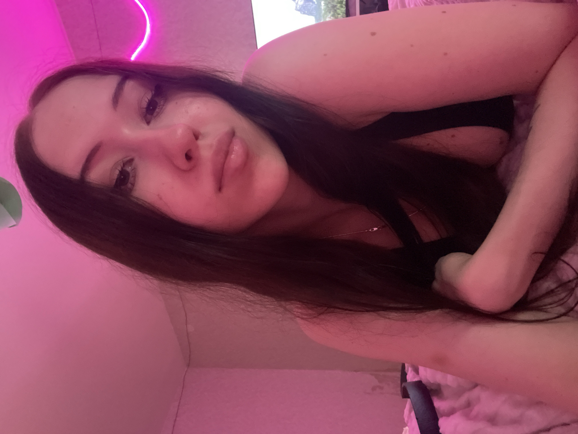 The image shows a young woman with long, dark hair lying down on a pink, fluffy surface, possibly a bed. She is wearing a black top. The lighting in the room is predominantly pink, with a neon pink light source visible in the background on the right side of the image. The background also includes a wall with some texture and a television screen displaying an image of mountains. To the left of the woman, a dark object, possibly a belt, is visible on the pink surface. There is a hint of a necklace on her chest. The image is a selfie, taken from a close-up perspective, focusing on the woman's face and upper body. The scene appears to be indoors, in a bedroom environment. No specific location in the city of Votkinsk, Russia can be determined from the image.