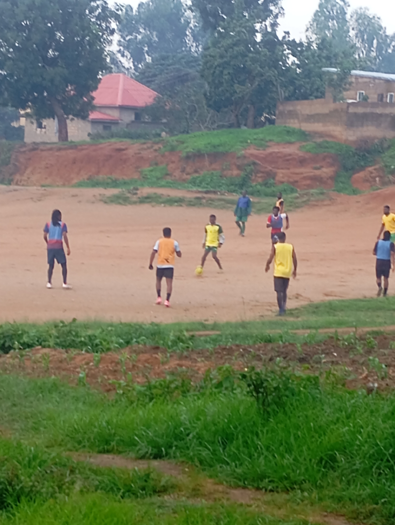 The image depicts an outdoor scene of individuals playing soccer on a dusty field. The location is identified as the city of Jos, Nigeria. Several young men are actively engaged in the game, with some running and others positioned on the field. One individual in the foreground is wearing a yellow vest and black shorts, appearing to be in motion. Another person in a blue and red shirt and dark shorts stands facing away from the camera. In the background, buildings with red roofs and brick walls are visible, suggesting a residential or semi-developed area. Trees and green vegetation border the field, and the ground is primarily composed of reddish-brown dirt. The overall atmosphere suggests a casual game of soccer taking place in a somewhat rural or less urbanized part of Jos.