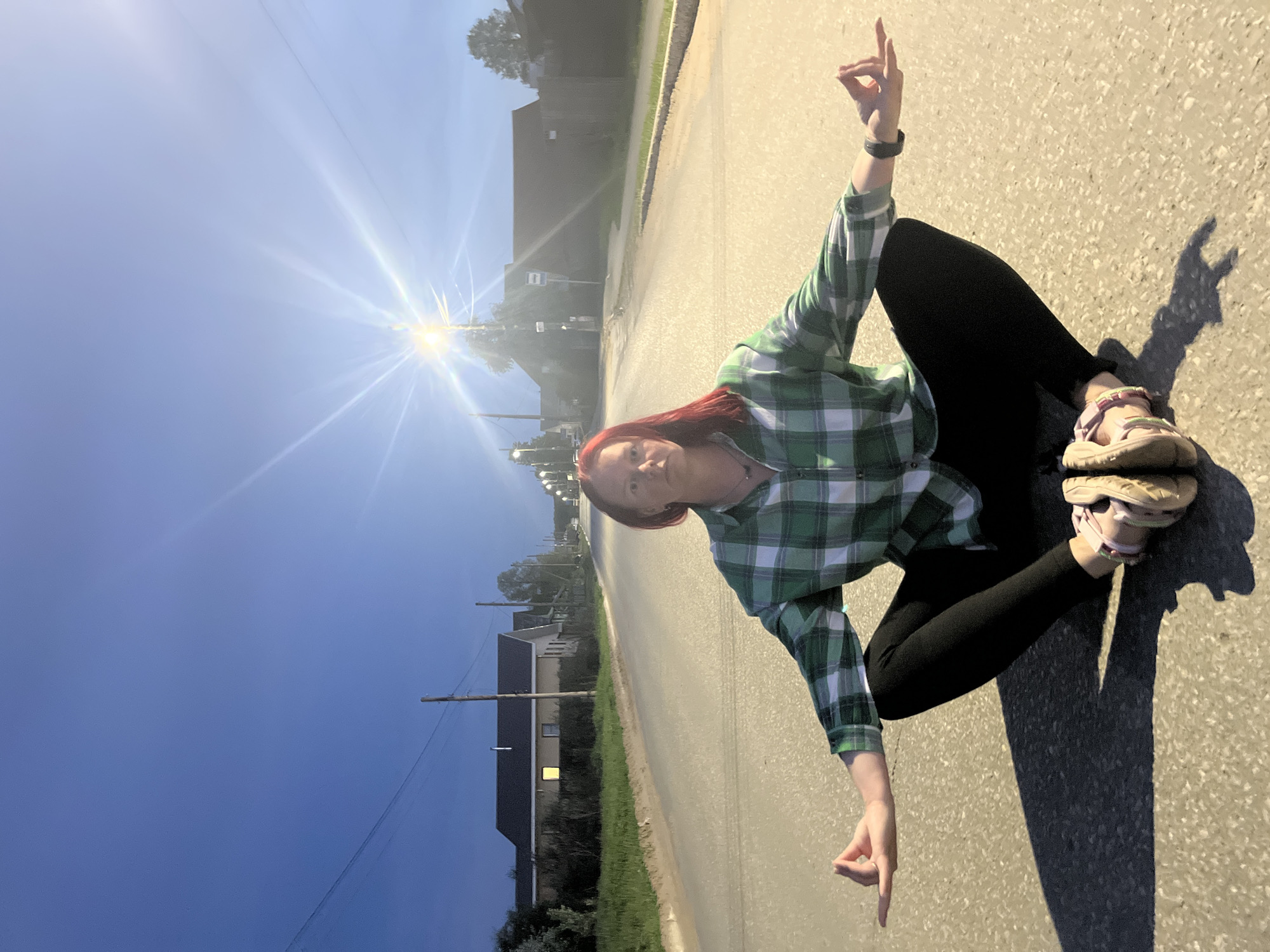 A woman sits cross-legged in the middle of an asphalt road. She wears a green and white plaid shirt, black leggings, and open-toed sandals. Her hair is red. She holds one hand up with her thumb and index finger touching. The other hand points to the ground. A long shadow is cast to her right. The road stretches into the distance with buildings and trees lining the sides. Overhead power lines are visible. A bright light illuminates the scene further down the road. The sky is blue. The location is the city of Chaikovsky, Russia.