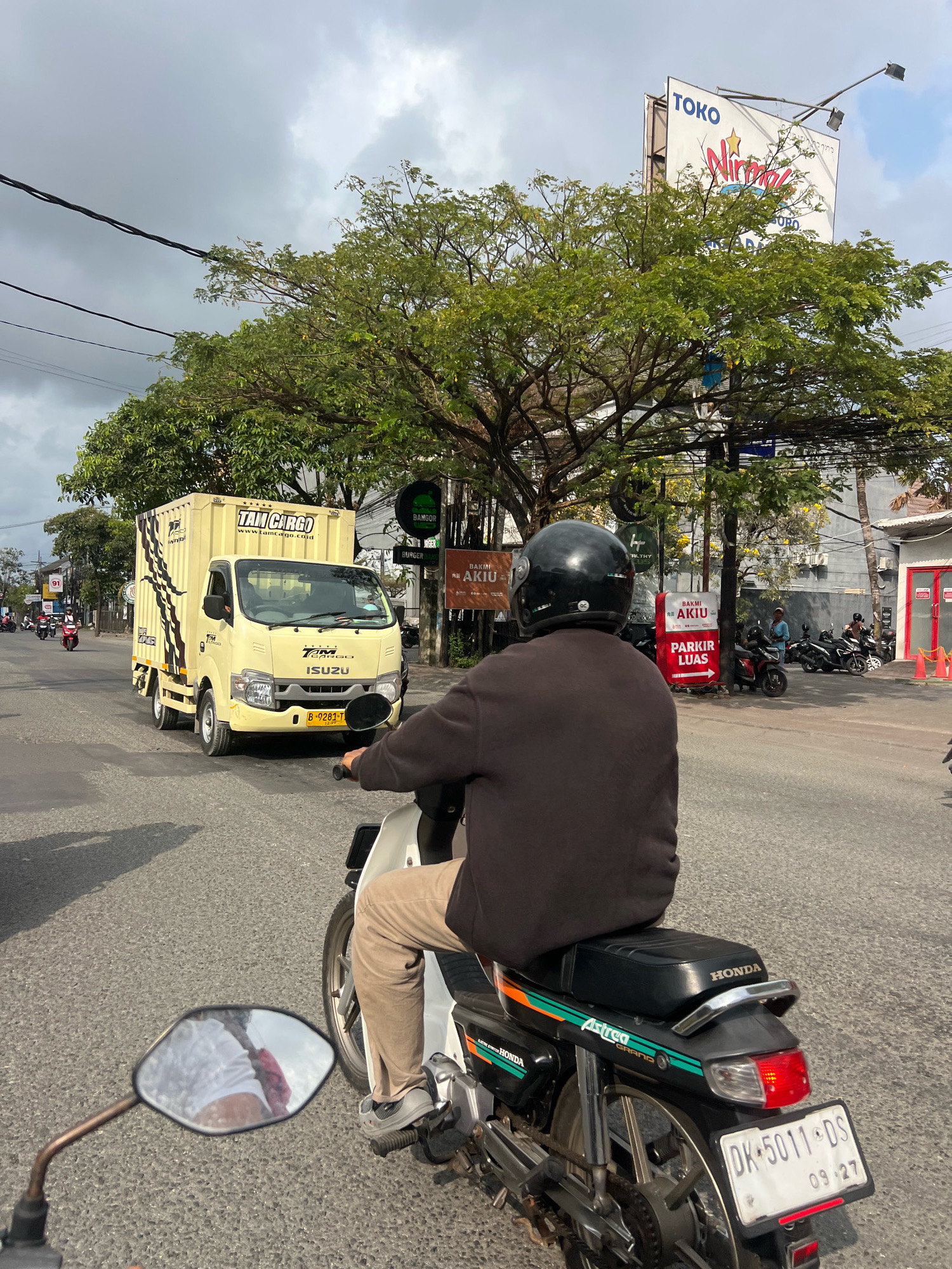 The image shows a street scene in Denpasar, Indonesia, on a cloudy day. In the foreground, a person on a black Honda Astrea Grand motorcycle is looking forward, with their back to the camera. The motorcycle has a license plate that reads "DK 5011 DS 09 27". The rider is wearing a black helmet and a dark brown long-sleeved shirt with tan pants and grey sneakers. A large yellow Isuzu truck with "TAM CARGO" written on its side is in the middle of the road ahead of the motorcycle. Other motorcycles and people are visible in the background, along with various businesses and billboards. The environment appears to be a busy urban area with trees and power lines.