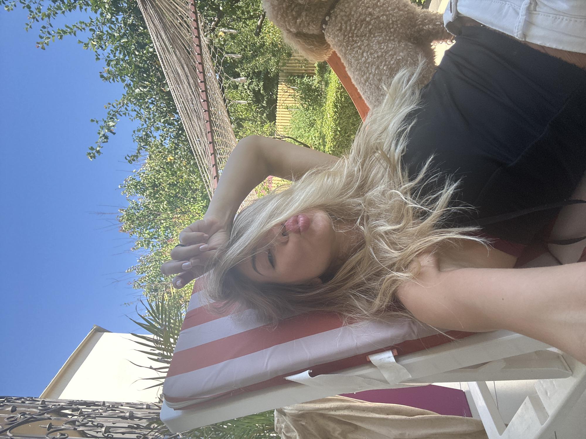 A blonde woman lies on a striped lounge chair in an outdoor setting, making a kissing face with her fingers near her face. She is wearing a black top and white shorts. A small brown poodle sits next to her. The background shows lush green trees and a clear blue sky. The presence of a building and a garden suggests a residential area, possibly within the city limits of Armavir, Russia, though the specific location cannot be definitively confirmed from the image alone.