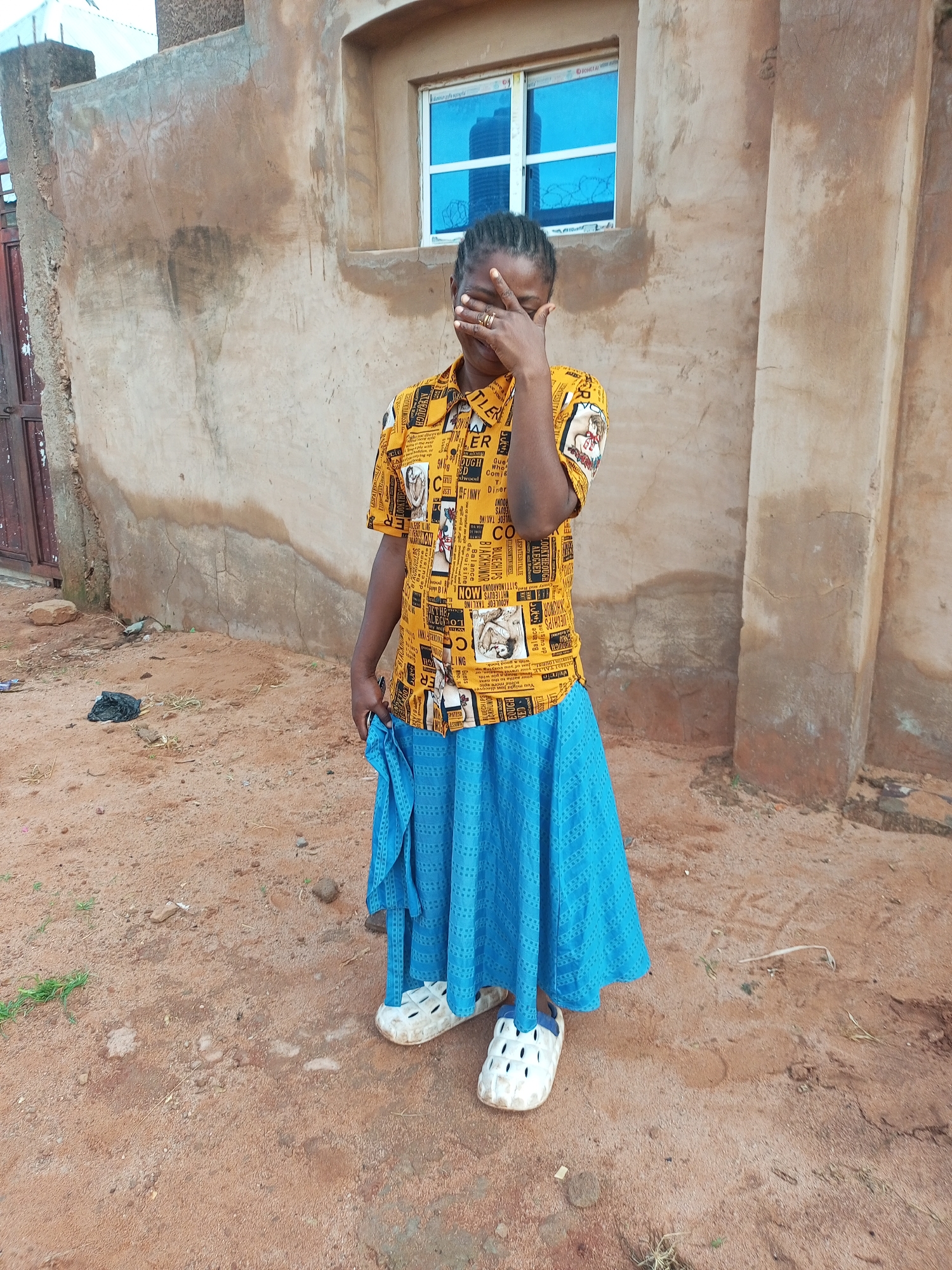 A young Black female, wearing a yellow patterned shirt and a blue skirt, stands outside a weathered, tan-colored building. The building has a small, blue-paned window and rough textured walls. The ground is covered in reddish-brown dirt and scattered debris. The female is covering her face with her right hand and wearing white perforated clogs. She is holding a blue garment in her left hand. The scene appears to be in an outdoor, undeveloped area, possibly a rural or informal settlement.