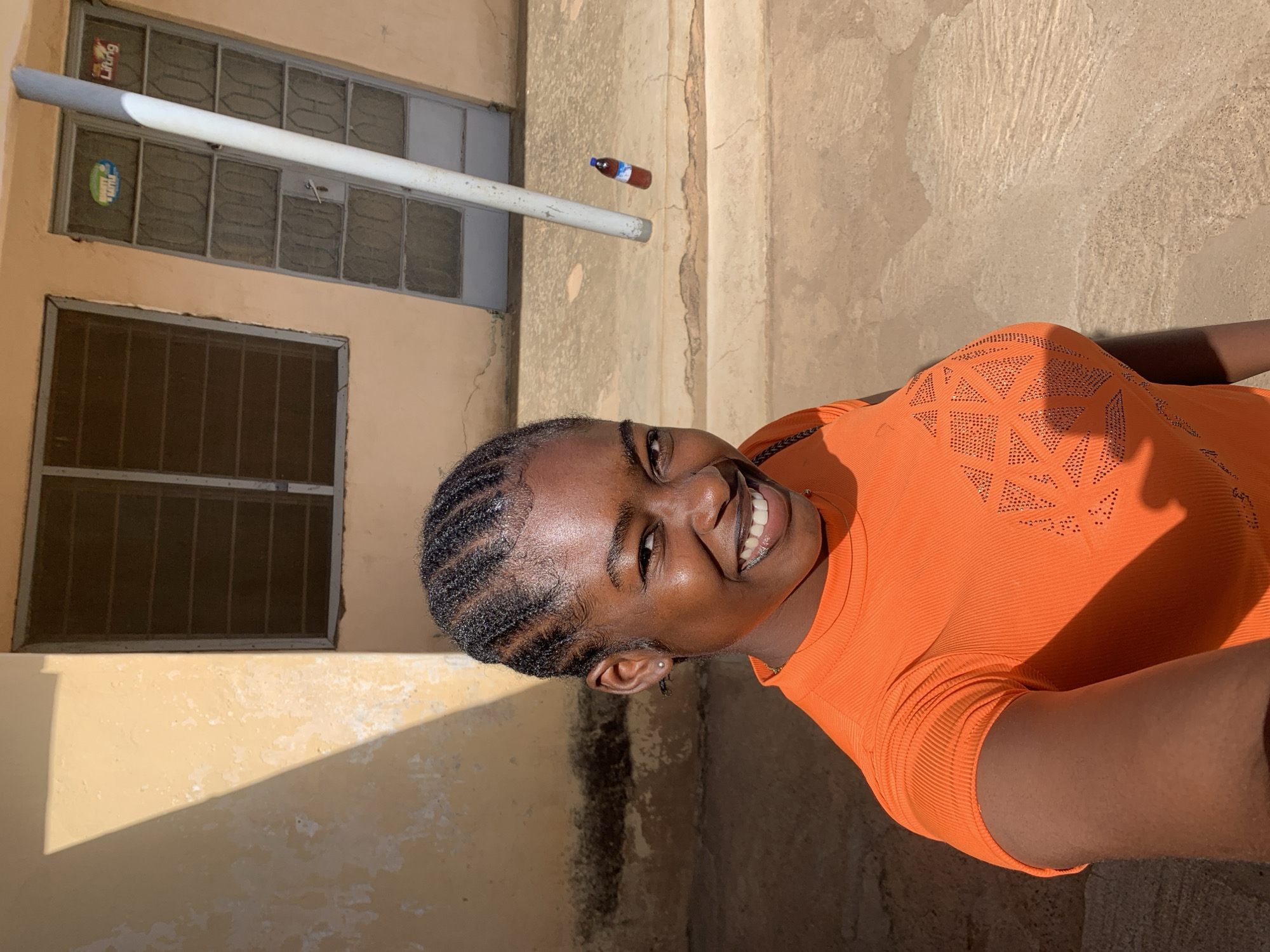 A young Black woman with braided hair and wearing an orange shirt is smiling at the camera. She is standing in front of a building, with a window visible behind her. Above the window is a pipe and a small bottle. The location appears to be outdoors, with a textured surface in the background. The photograph was taken in the city of Rafin Par, Nigeria.