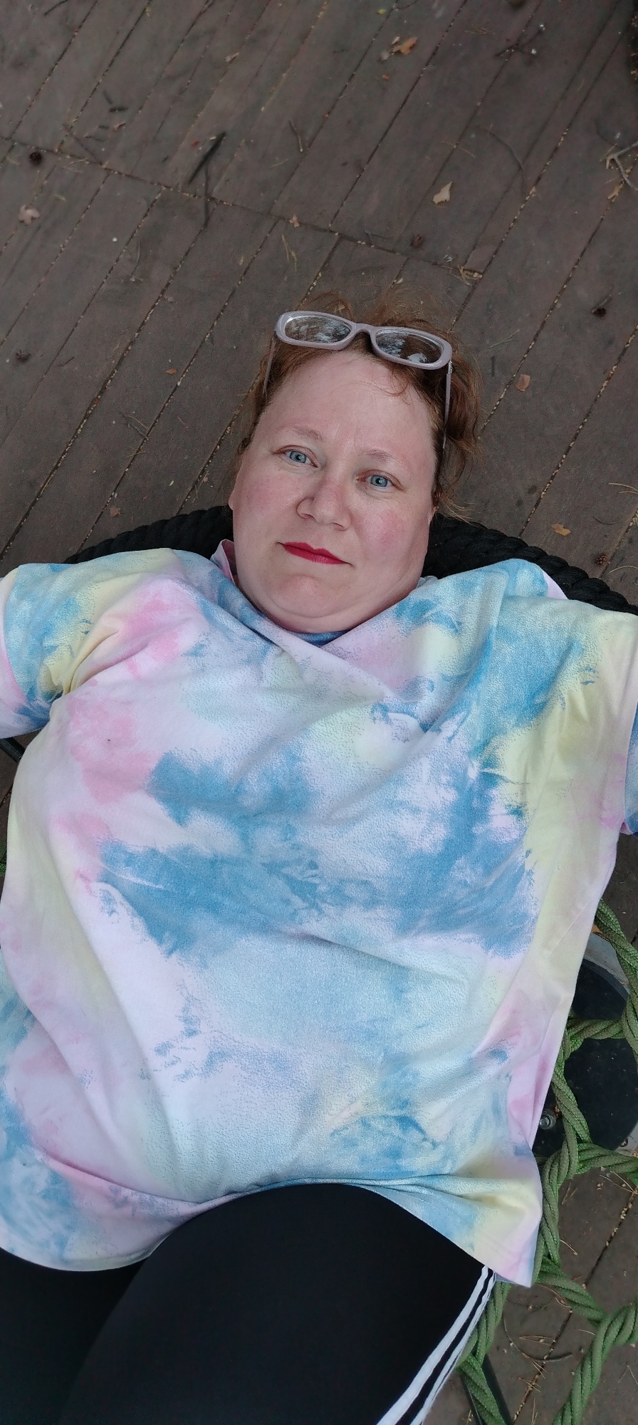 A woman with reddish hair, wearing glasses on her head, lies in a black rope swing. She wears a tie-dye shirt in pastel colors and black leggings with a white stripe down the side. The swing is situated on a wooden deck. There is no specific indication of Chaikovsky, Russia.