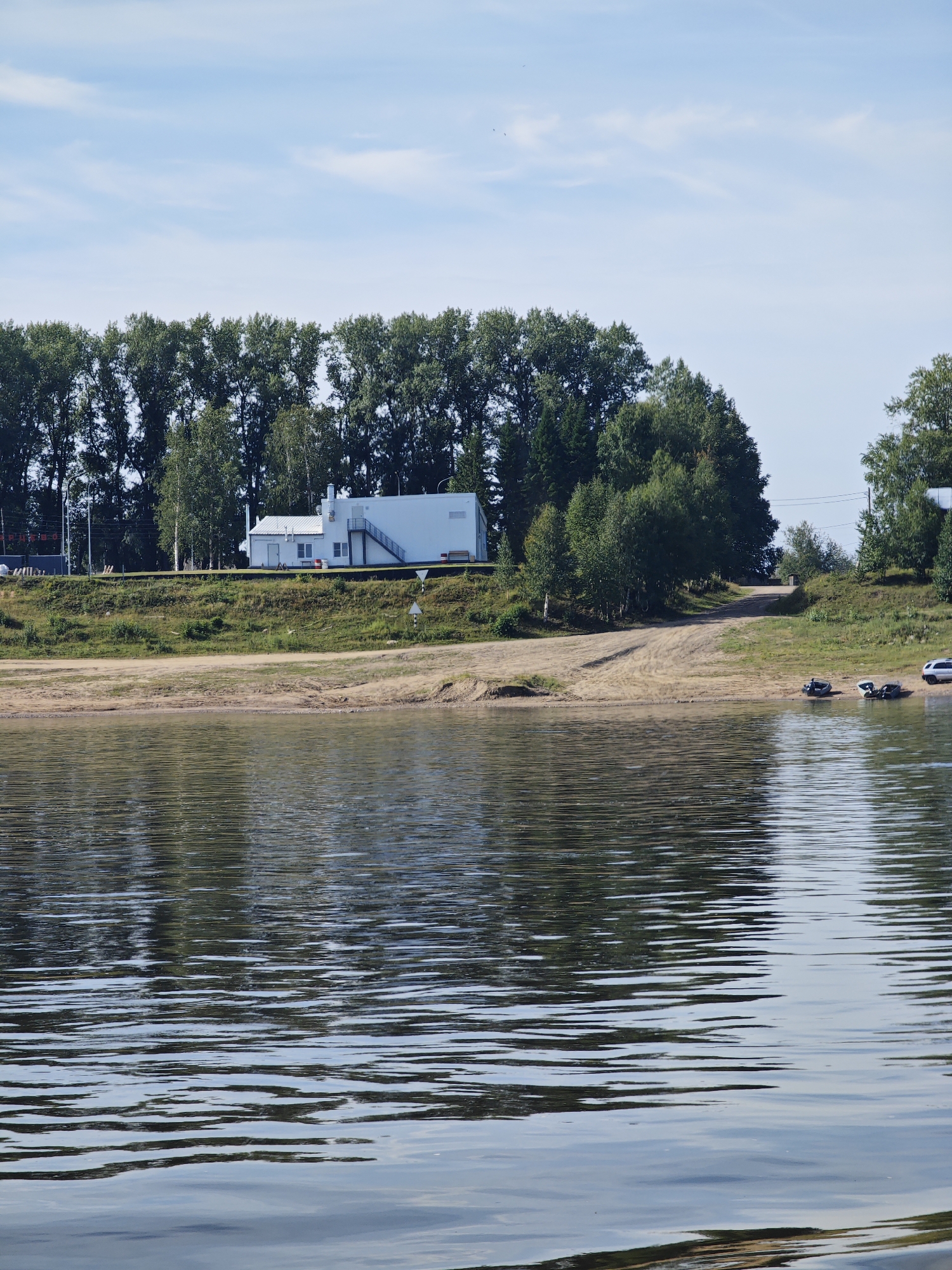 A wide, calm river occupies the foreground, reflecting the trees and sky. The middle ground features a sandy embankment leading up to a grassy hill. On the hill, a modern white building with a prominent staircase is situated amongst a dense line of trees. Several boats are pulled up onto the sandy bank, and a silver car is parked on the right side of the embankment. The background is dominated by the lush green foliage of trees under a clear blue sky with scattered clouds. The scene appears to be a tranquil riverside location, potentially a recreational area or the grounds of a riverside facility.