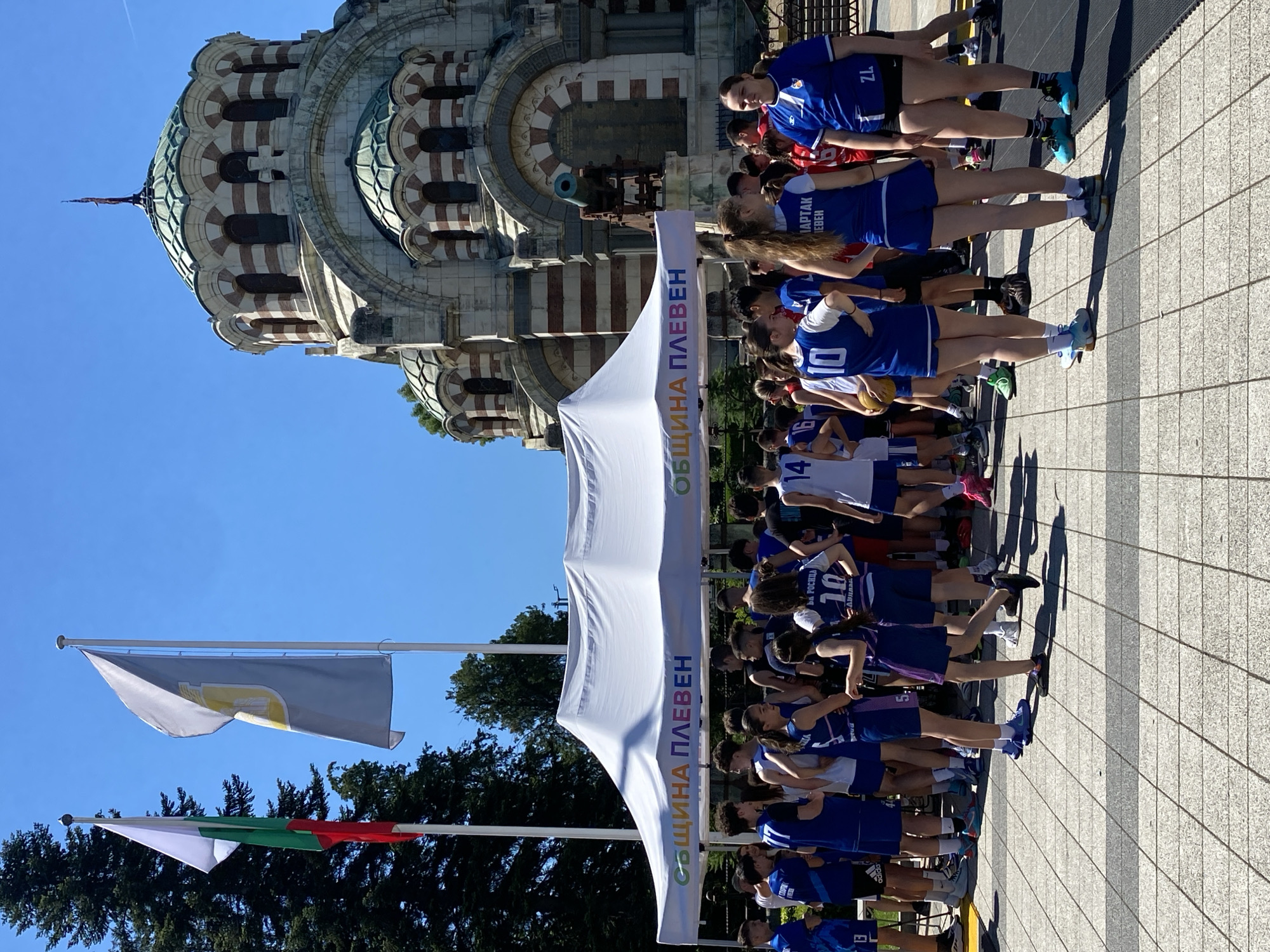 The image captures a scene outside the St. George the Conqueror Rotunda in Pleven, Bulgaria, on a sunny day. A group of young people, predominantly in blue and white basketball uniforms, are gathered around a white tent. The tent bears the inscription "ОБЩИНА ПЛЕВЕН" (Municipality of Pleven) in colorful letters. Some individuals are holding a basketball. In the background, the distinct architecture of the Rotunda, with its copper dome and striped stonework, is visible against a clear blue sky. Two flags, one the Bulgarian national flag and another with a gray background and a yellow emblem, fly from poles near the Rotunda. The ground is paved with patterned tiles. The atmosphere appears to be that of an outdoor event or sporting activity.