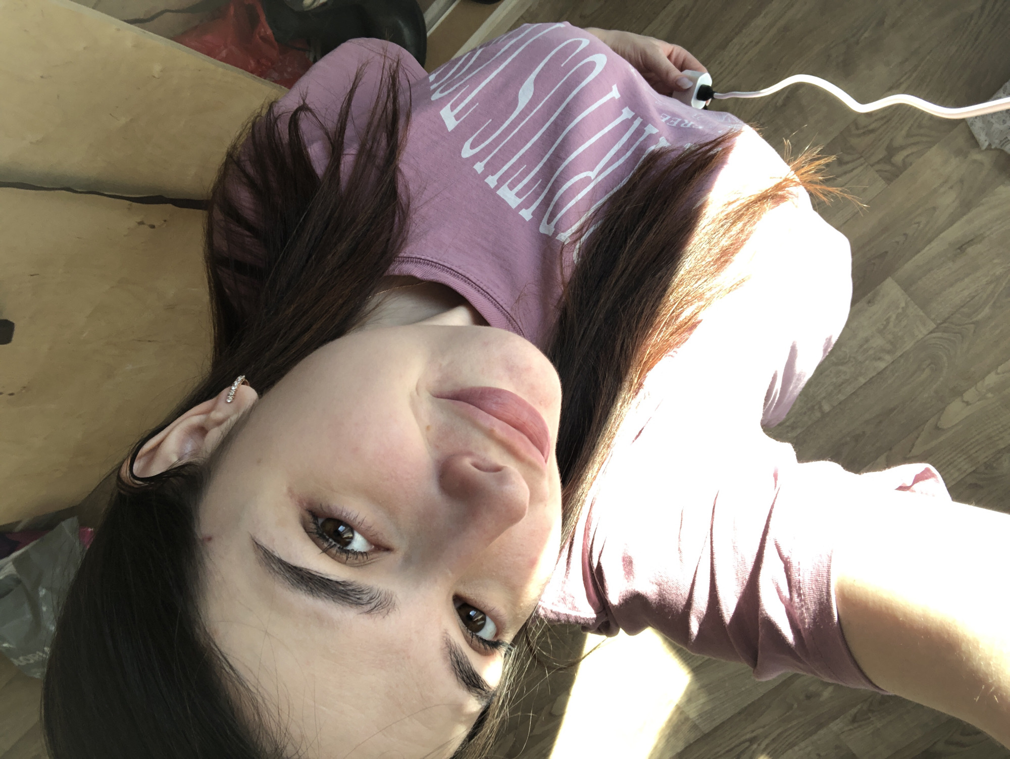 A woman is lying on a wooden floor, wearing a pink long-sleeved shirt. Her dark brown hair frames her face, and she has an earring in her left ear. A white electrical cord is visible near her shoulder. The background includes a partial view of a staircase and some unidentified objects. The location cannot be determined from the image.