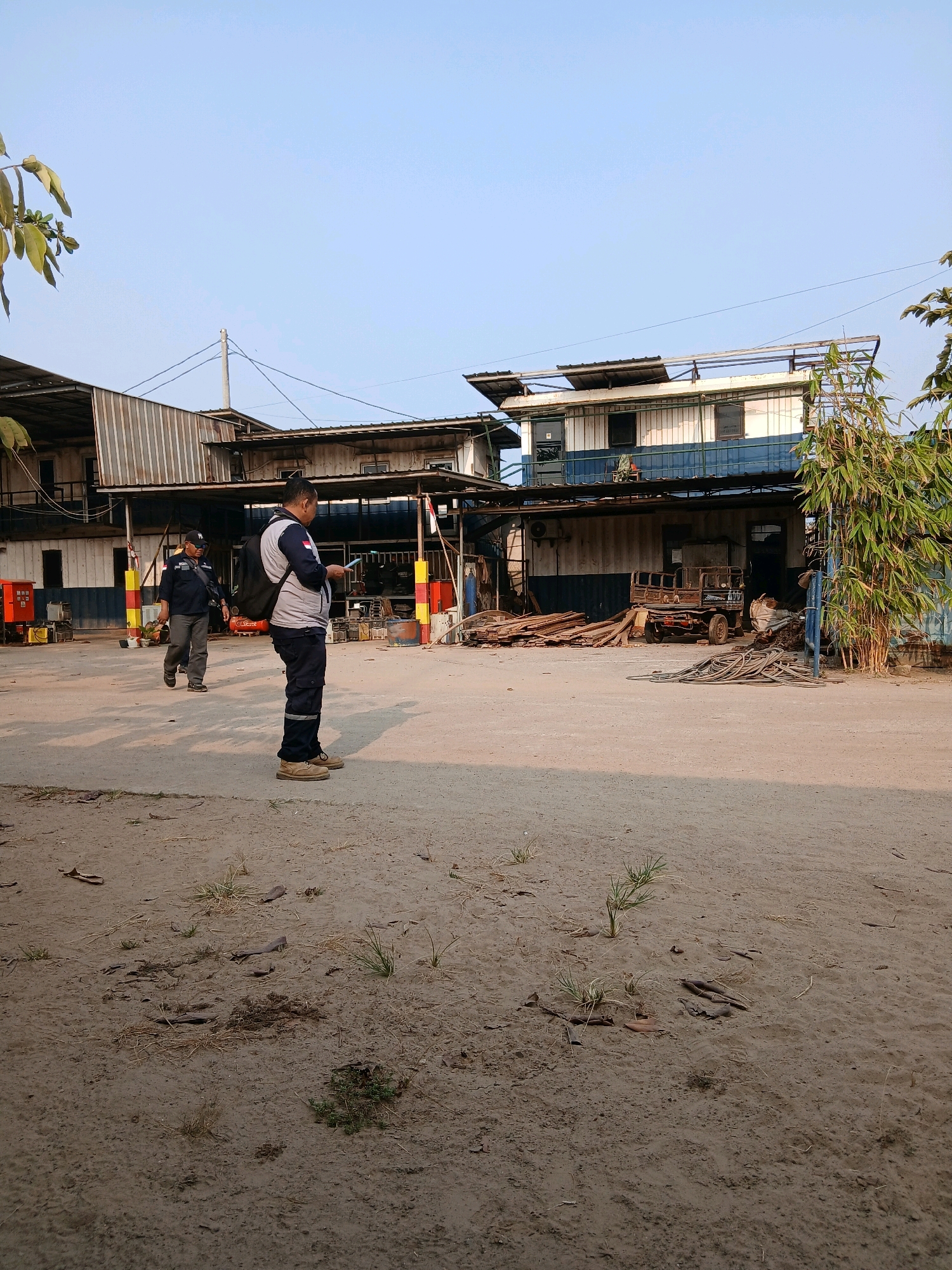 The image shows an outdoor industrial or workshop area in Pangkalpinang, Indonesia. Two men are visible. The man in the foreground is wearing a blue and grey work uniform, dark pants, and tan work boots, and he is looking at his phone while holding a tablet. He has a black backpack. The second man, in the background and walking to the left, is wearing a dark blue uniform, dark pants, and a black baseball cap.
The environment includes a dirt and gravel ground with scattered debris. In the background, there are several buildings with corrugated metal roofs. One building is two stories high with a blue and white facade. There are piles of wood, metal pipes, a small rusty truck, and various pieces of industrial equipment. Bamboo plants are growing to the right of the frame. The sky is clear and blue, indicating daylight. The lighting suggests it is late afternoon.