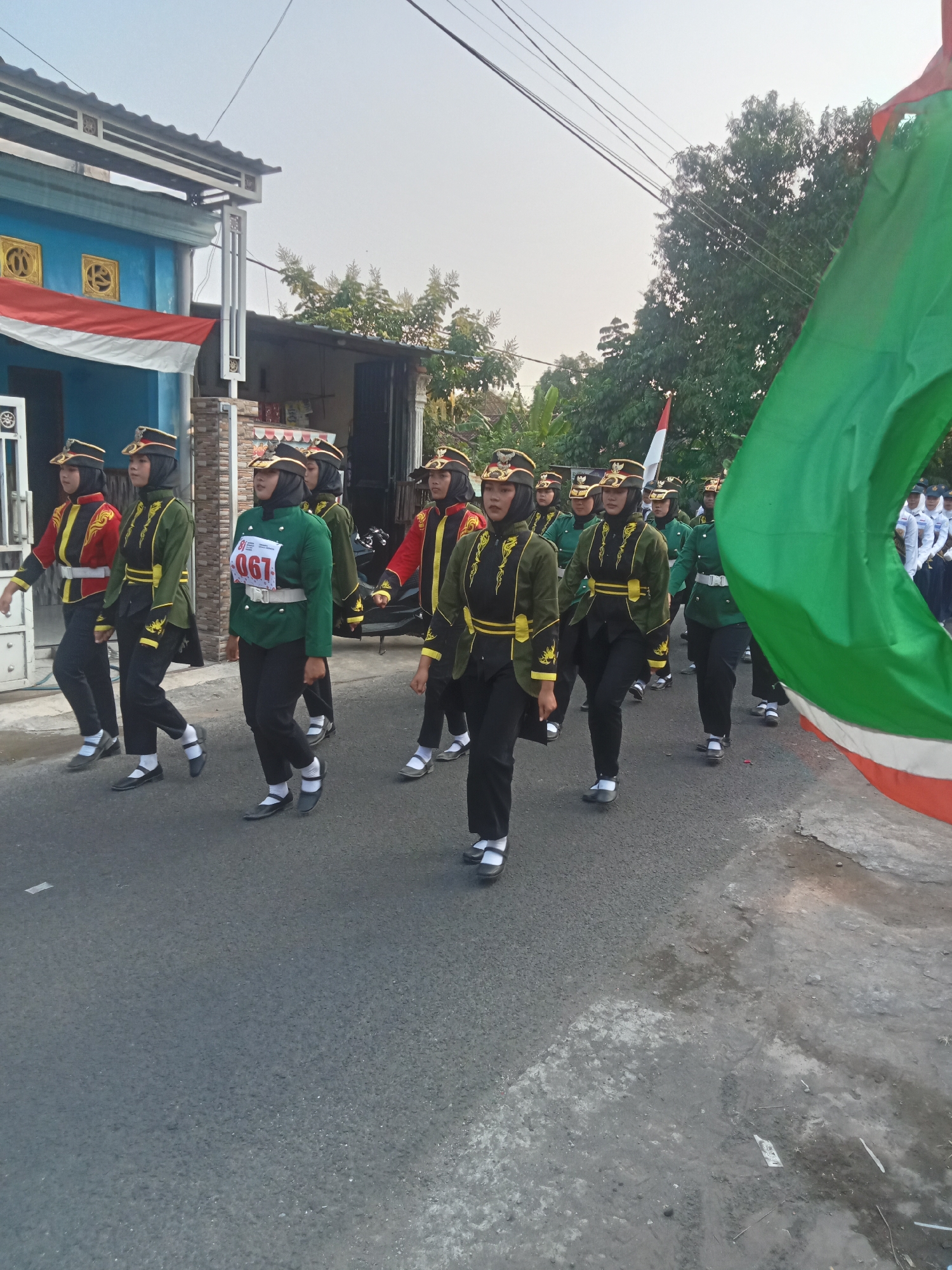 In an outdoor, street-level scene, a group of young people in uniform march. The uniforms consist of dark trousers, black shoes with white socks, and a variety of jackets in green and red. The jackets have yellow trim and some are adorned with gold embroidery. Many of the marchers wear black headscarves under their hats, which are dark with a gold emblem. One individual wears a green jacket with a bib on the front displaying the number "067" in red. A large green and red flag is prominently displayed in the foreground, partially obscuring the view of the marchers. The background shows buildings with blue and white facades, trees, and a clear sky. The ground is asphalt, with some debris visible.