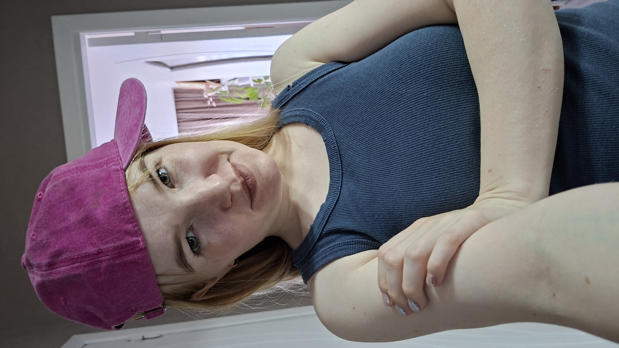 A fair-skinned woman with freckles and blonde hair is photographed from a low angle, wearing a dark blue ribbed tank top and a pink velvet baseball cap. The cap is positioned so that the brim is partially obscuring her forehead and left eyebrow. Her arms are crossed, and she is looking directly at the camera with a slight smile. Behind her is a white doorway or window frame, with a partially visible window and what appears to be indoor plants in the background. No specific details indicate the location is Chaikovsky, Russia.