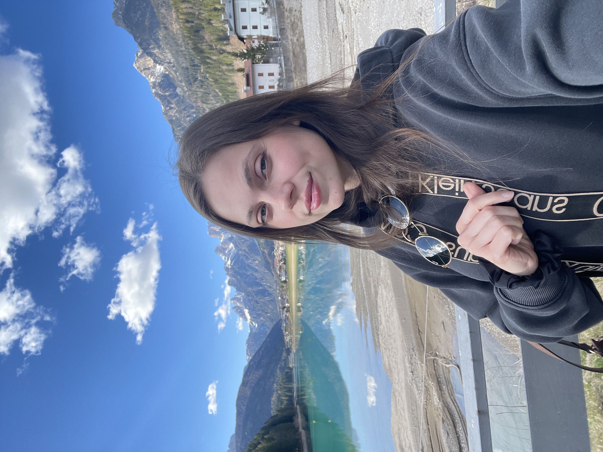 A young woman with long brown hair takes a selfie in Auronzo di Cadore, Italy. She is wearing a black hooded sweatshirt with a Calvin Klein logo band around the neckline. Her sunglasses are casually hooked onto the neckline of her shirt. She holds a black scrunchie in her right hand. Behind her is a picturesque view of the Tre Cime di Lavaredo mountains reflected in the blue-green waters of Lake Auronzo. White clouds dot the bright blue sky. Buildings can be seen lining the shore of the lake, and the surrounding area appears to be a mix of sand and greenery. The woman is leaning against a gray metal railing.