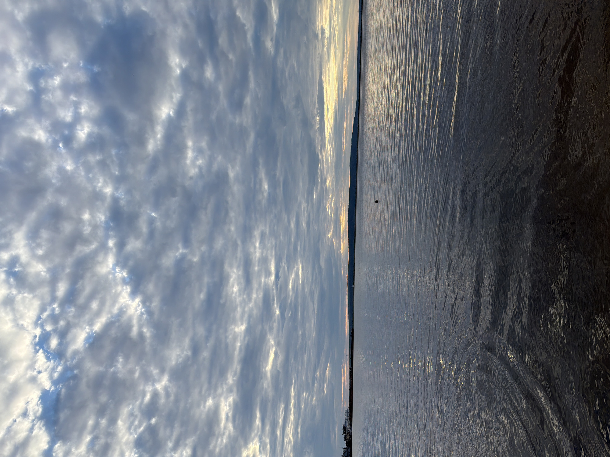 This image captures a wide, calm body of water, likely a river or a large lake, under a cloudy sky. The water surface is rippled, reflecting the muted light from the overcast sky. In the distance, a low-lying landmass with a dark silhouette forms the horizon. On the left side of the horizon, there's a cluster of structures, possibly industrial buildings or port facilities, with a few white vessels docked. The sky is filled with a textured pattern of gray and white clouds, with patches of brighter light peeking through, suggesting either sunrise or sunset. The overall atmosphere is serene and somewhat somber due to the cloudy conditions. The location is identified as Chaikovsky, Russia, implying this is a view of the Kama River or a reservoir within the city's vicinity.