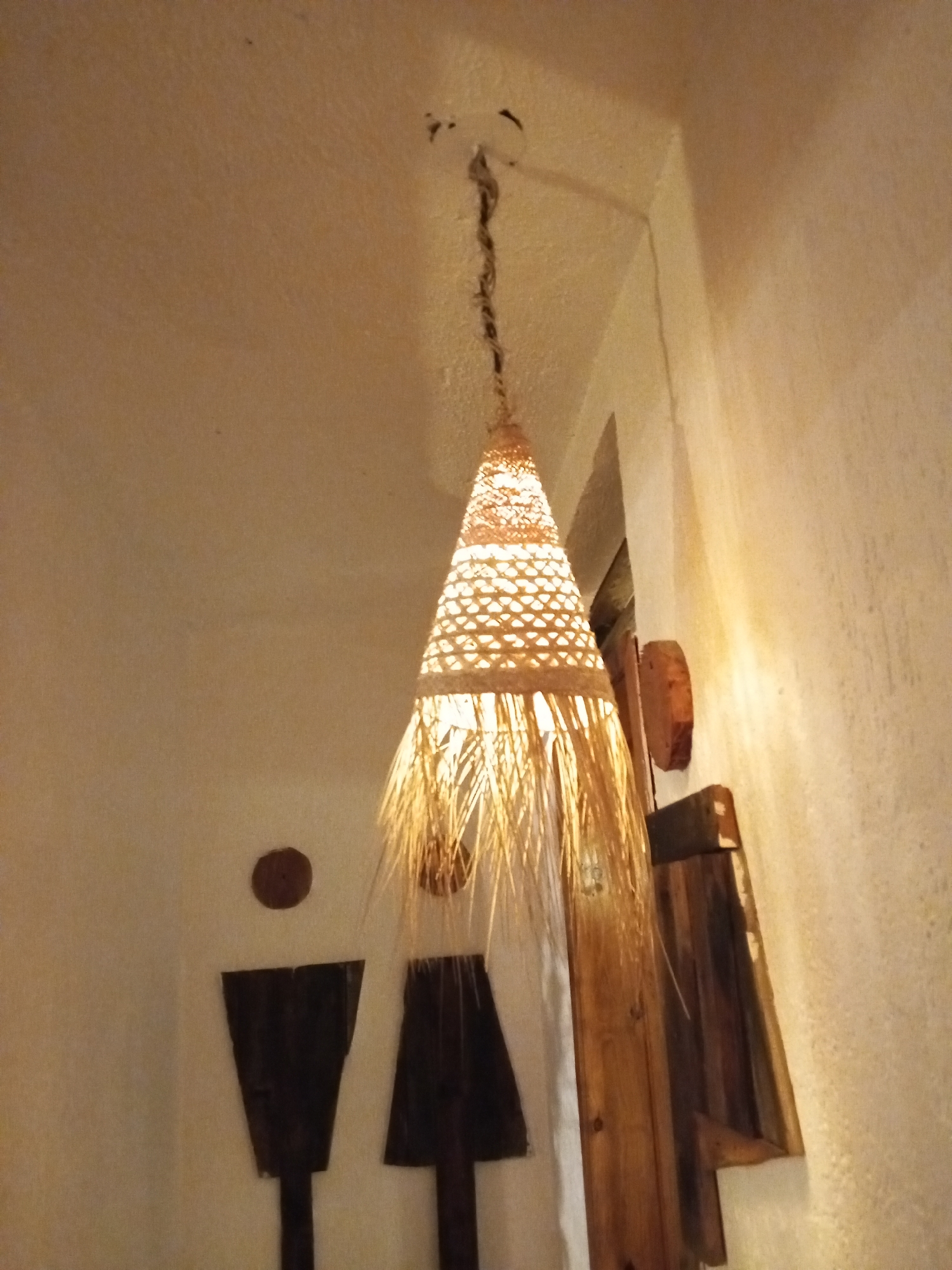 The image shows a dimly lit interior, likely of a restaurant or bar, possibly in Porlamar, Venezuela. A cone-shaped woven lamp with straw-like fringe hangs from the ceiling. On the wall are dark, stylized restroom signs representing male and female figures, and a partially visible wooden structure. The walls are textured and painted in warm, muted tones. The lighting is soft and creates shadows.