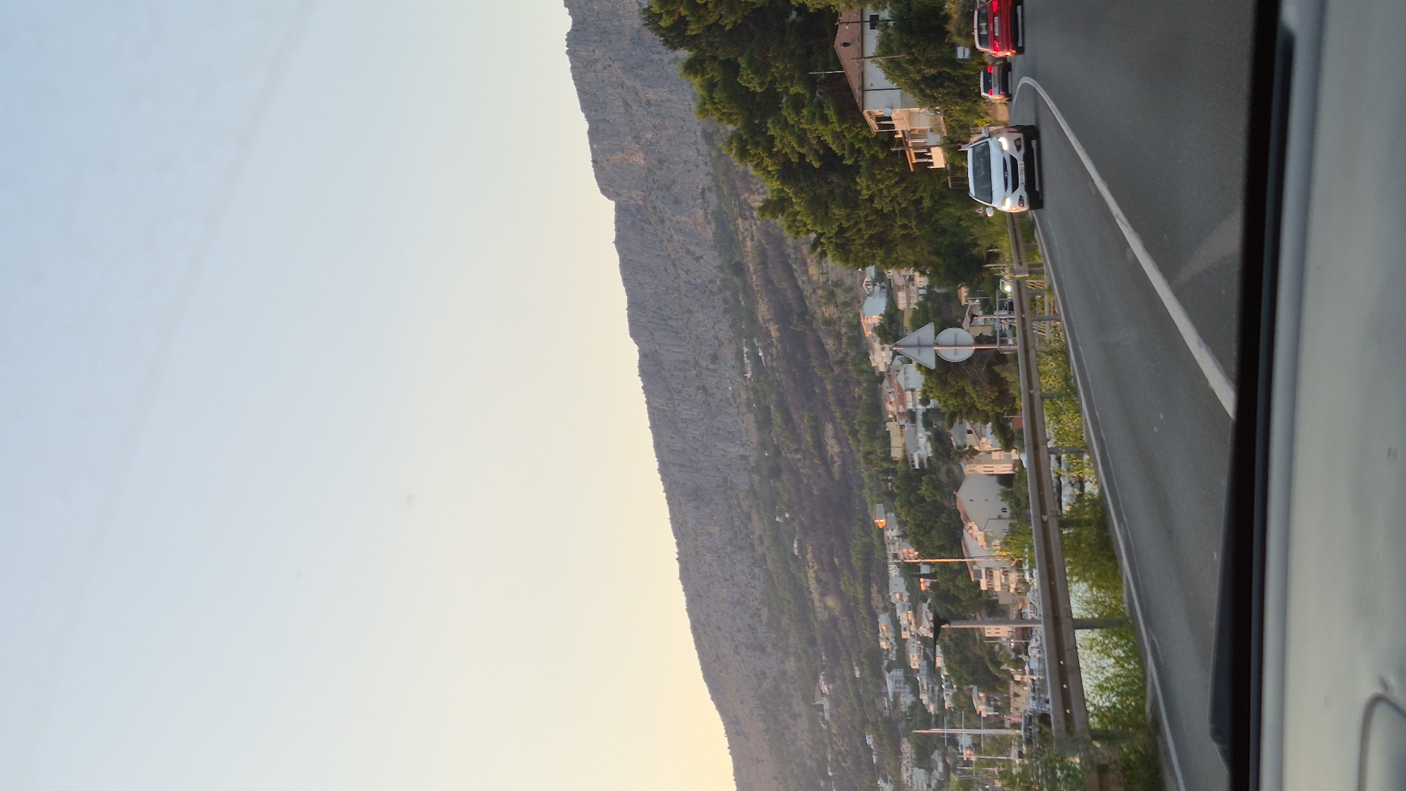 An eye-level, medium shot captures a view from a car traveling along a coastal road. The road curves to the right, with a guardrail separating it from a body of water and a town. The town is nestled against a steep, rocky mountainside covered in sparse vegetation and dotted with white buildings. Green trees, some with dark patches suggesting a recent fire, line the road and the lower slopes of the mountain. Several cars are visible on the road; a white SUV is in the foreground, followed by a red sedan. The sky is clear and pale blue, transitioning to a soft yellow near the horizon. The overall impression is a scenic drive along a Mediterranean coast during the late afternoon or early evening.