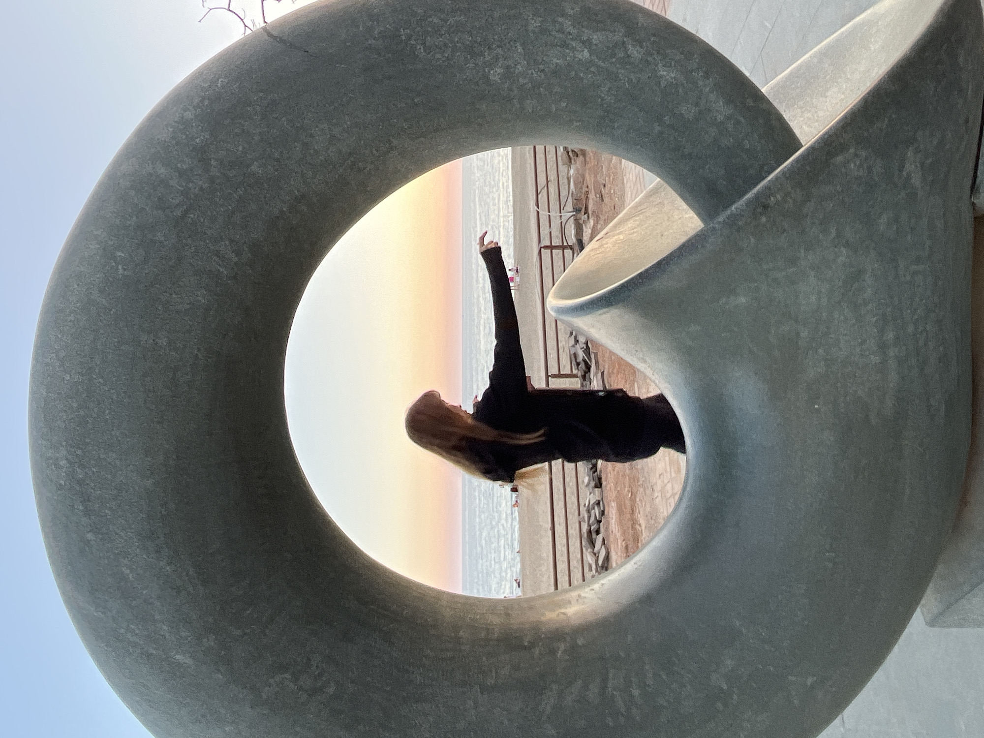 A vertically oriented photograph frames a woman and a serene seascape through a large, abstract sculpture. The sculpture, likely made of concrete or stone, is a thick, grey, loop-like structure, possibly resembling intertwined rings or a Mobius strip, dominating the foreground. A woman with long blonde hair, dressed in dark attire, is positioned within the central opening of the sculpture. She stands with her back mostly to the viewer, gazing towards the horizon, with her left arm raised and hand extended upwards, as if reaching out or embracing the view.
Beyond the sculpture, a calm body of water, indicative of the sea or ocean, extends into the distance. The sky above the water is painted with soft, diffused hues of pale yellow, orange, and pink, characteristic of either sunrise or sunset. A narrow strip of what appears to be a shoreline or rocky beach separates the water from the immediate background behind the woman. The overall atmosphere is tranquil and contemplative. No text is visible in the image. This scene is located in Alanya, Turkey.