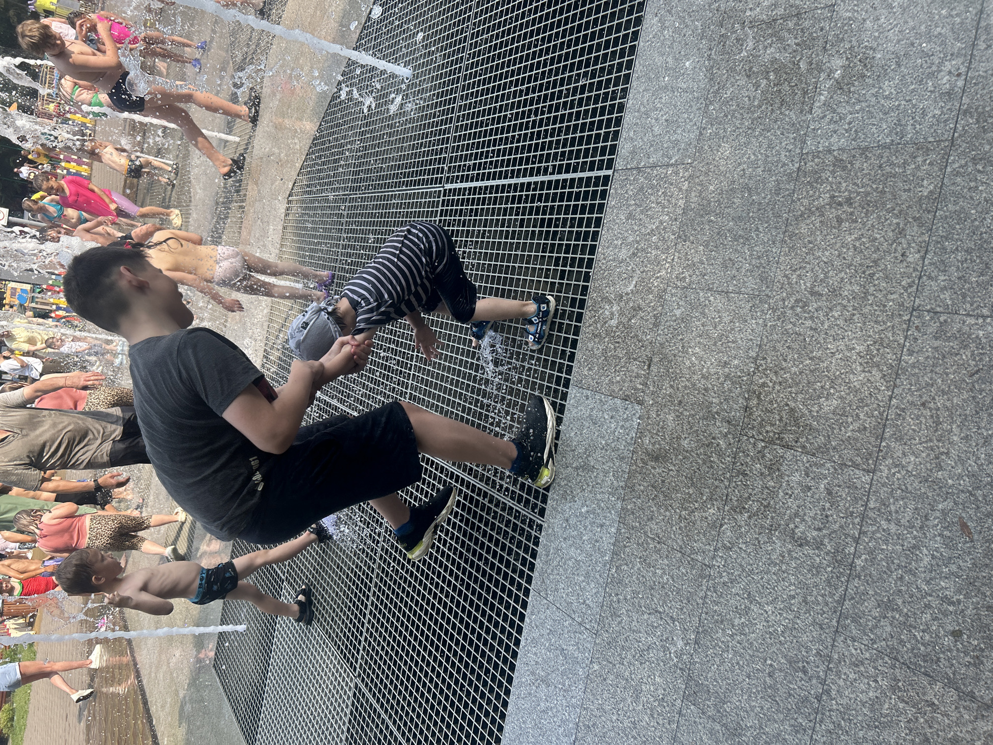 In a public splash pad in Omsk, Russia, two boys are playing in the water. The older boy, wearing black shorts, is standing and appears to be helping the younger boy. The younger boy, in a striped t-shirt and black shorts, is crouching down and interacting with the water. Other children and adults are visible in the background, enjoying the water park. The ground is covered with a metal grate and granite paving stones.