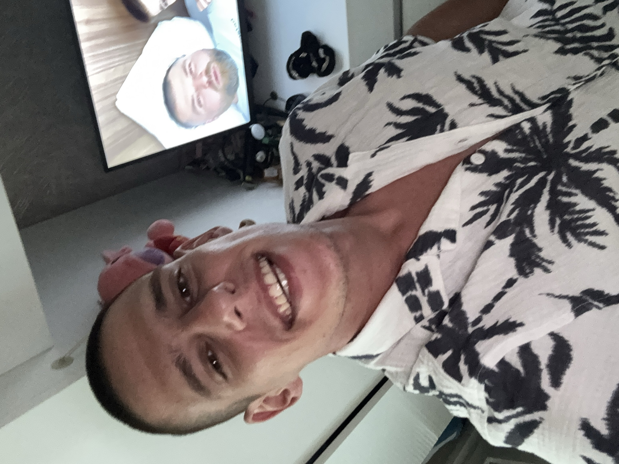 A man is taking a selfie. He has short, dark hair and is wearing a white shirt with a black tropical print. He is smiling and looking at the camera. In the background, there is a television screen showing another man. There is also some furniture and decorations visible in the background. It is not possible to determine the location in the city of Sofia, Bulgaria from the image.