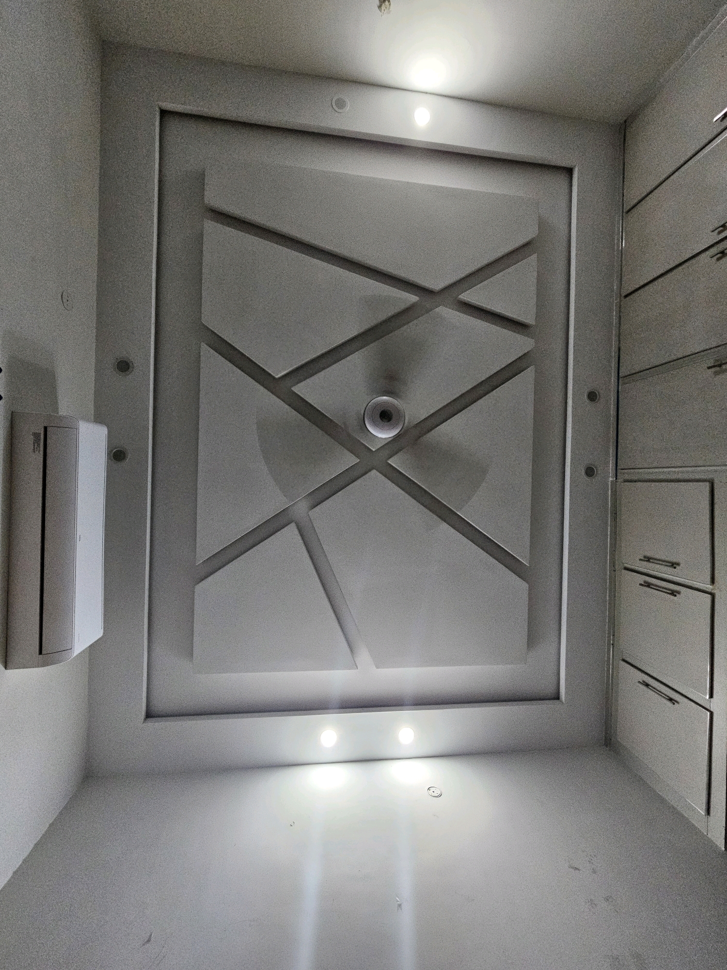 The image shows a modern room with a geometric coffered ceiling and built-in cabinetry. The ceiling features recessed lighting and a central circular fixture, possibly a fan or light. The walls are painted a neutral color, and an air conditioning unit is mounted on the left wall. The cabinetry on the right appears to be floor-to-ceiling with a sleek, handle-less design and subtle horizontal accents. The overall aesthetic is clean and contemporary. The image is a photograph taken from a low angle looking upwards towards the ceiling. No people are visible in the image. The location is identified as Jawad Avenue, Pakistan.