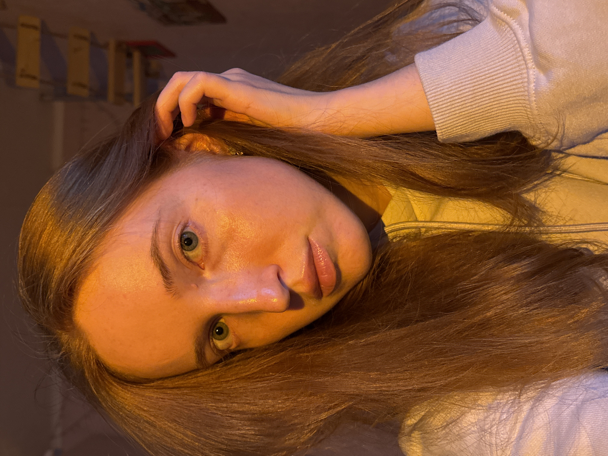 A close-up portrait features a young woman with long, reddish-brown hair, fair skin, and striking light green or blue-green eyes. She is positioned diagonally across the frame, looking directly at the viewer with a calm and engaging gaze. Her right hand is gently raised, with fingers lightly curled and resting near her temple, seemingly interacting with her hair. She is wearing a light-colored, possibly beige or off-white, ribbed sweatshirt or top. The image is lit with warm, artificial light, casting a glow on her face and hair, suggesting an indoor setting during the evening or night. The background is dimly lit and softly blurred, revealing indistinct shapes that hint at shelving or furniture in a room. No text is visible in the image. The scene is set in Chaikovsky, Russia.