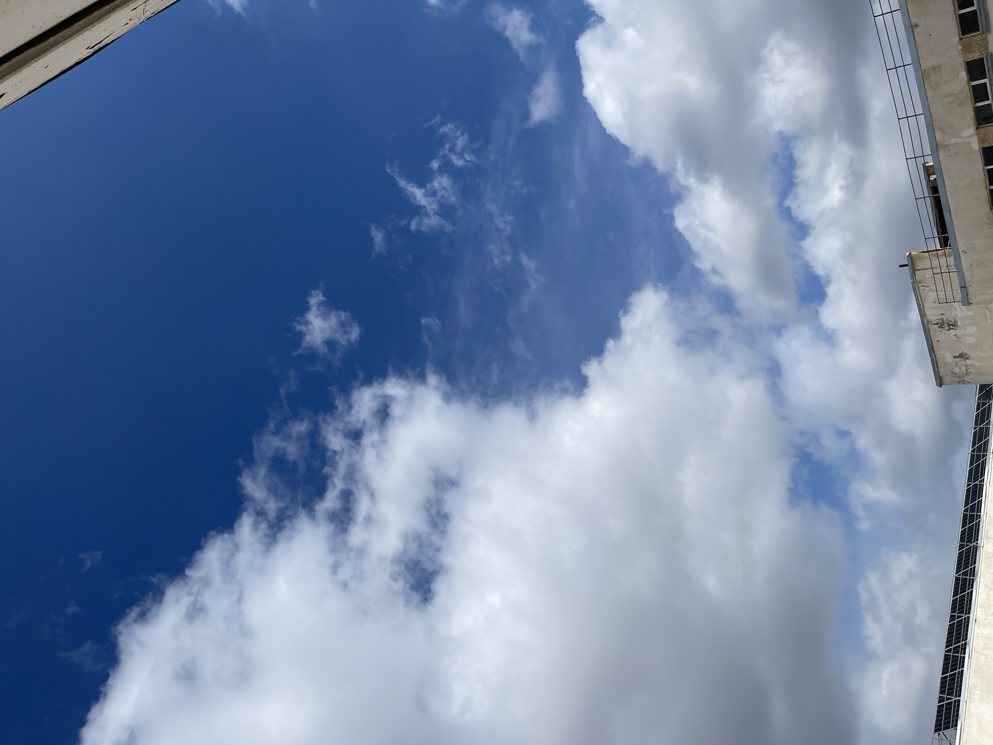 The image shows a view of the sky in Navoiy City, Uzbekistan, with a portion of a building visible in the upper right and upper left corners. The sky is primarily blue, interspersed with white clouds. The building appears to be multi-story, with visible windows, balconies, and a metallic railing. A solar panel array is affixed to the building on the right.