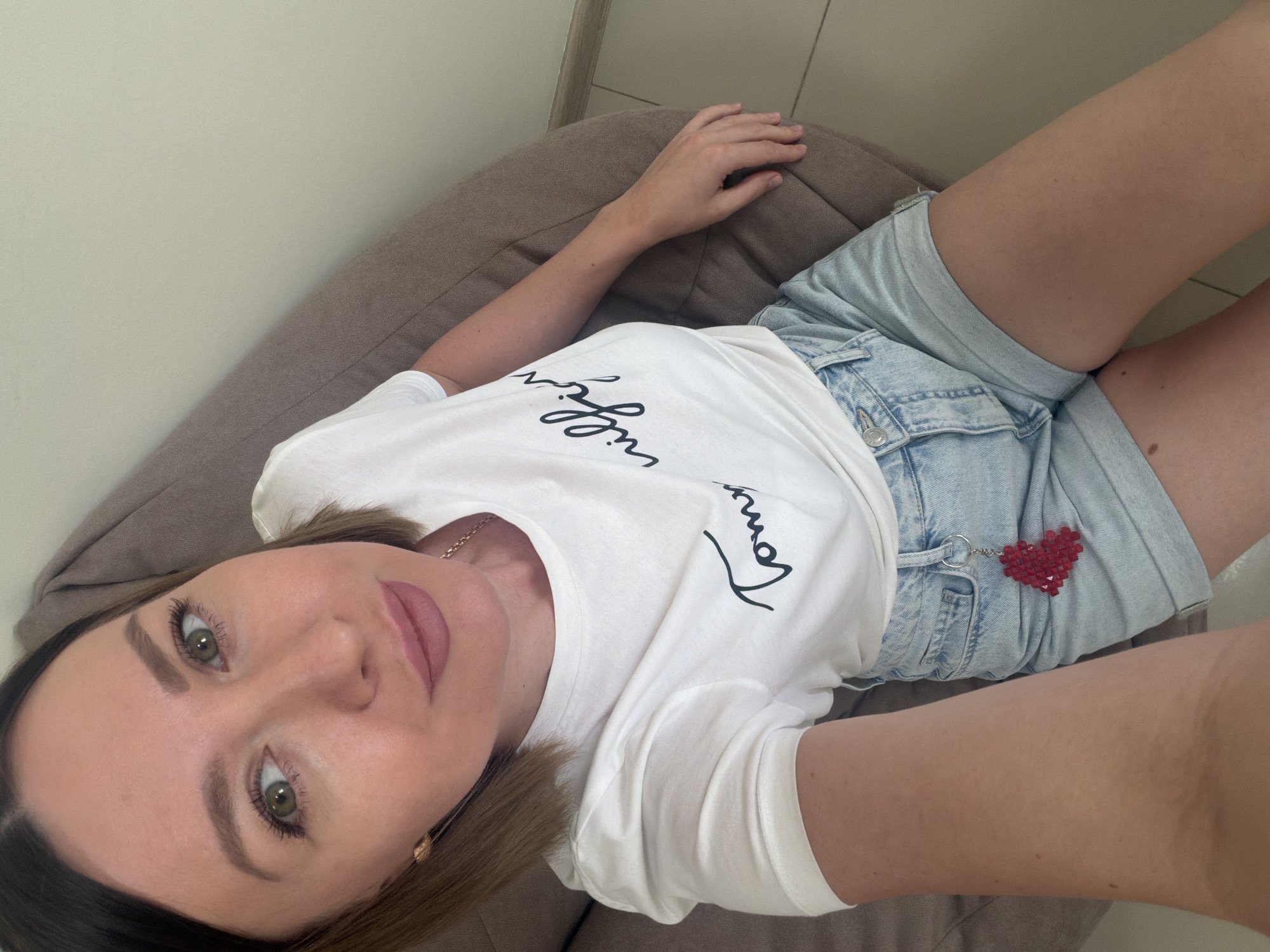 A woman is captured in a high-angle selfie, lying down on a light brown, possibly fabric or beanbag-like, surface. Her upper body and legs are visible, set against a plain, light-colored wall. She appears to be in her 20s or 30s, with light brown hair framing her face. She has green eyes, defined eyebrows, mascara, and a pinkish lip tint, gazing directly at the camera with a calm expression.
She is dressed in a white short-sleeved t-shirt featuring black script text across the chest. Parts of the text are visible, including what looks like "I'll" or "I will" and "Tommy". Below the t-shirt, she wears light blue denim shorts. A distinct red, heart-shaped beaded keychain is attached to the belt loop of her shorts. A delicate gold-colored chain necklace with a small pendant is also visible around her neck. The scene appears to be indoors with ample, soft lighting, suggesting it's daytime. This image was taken in Rostov-on-Don, Russia.