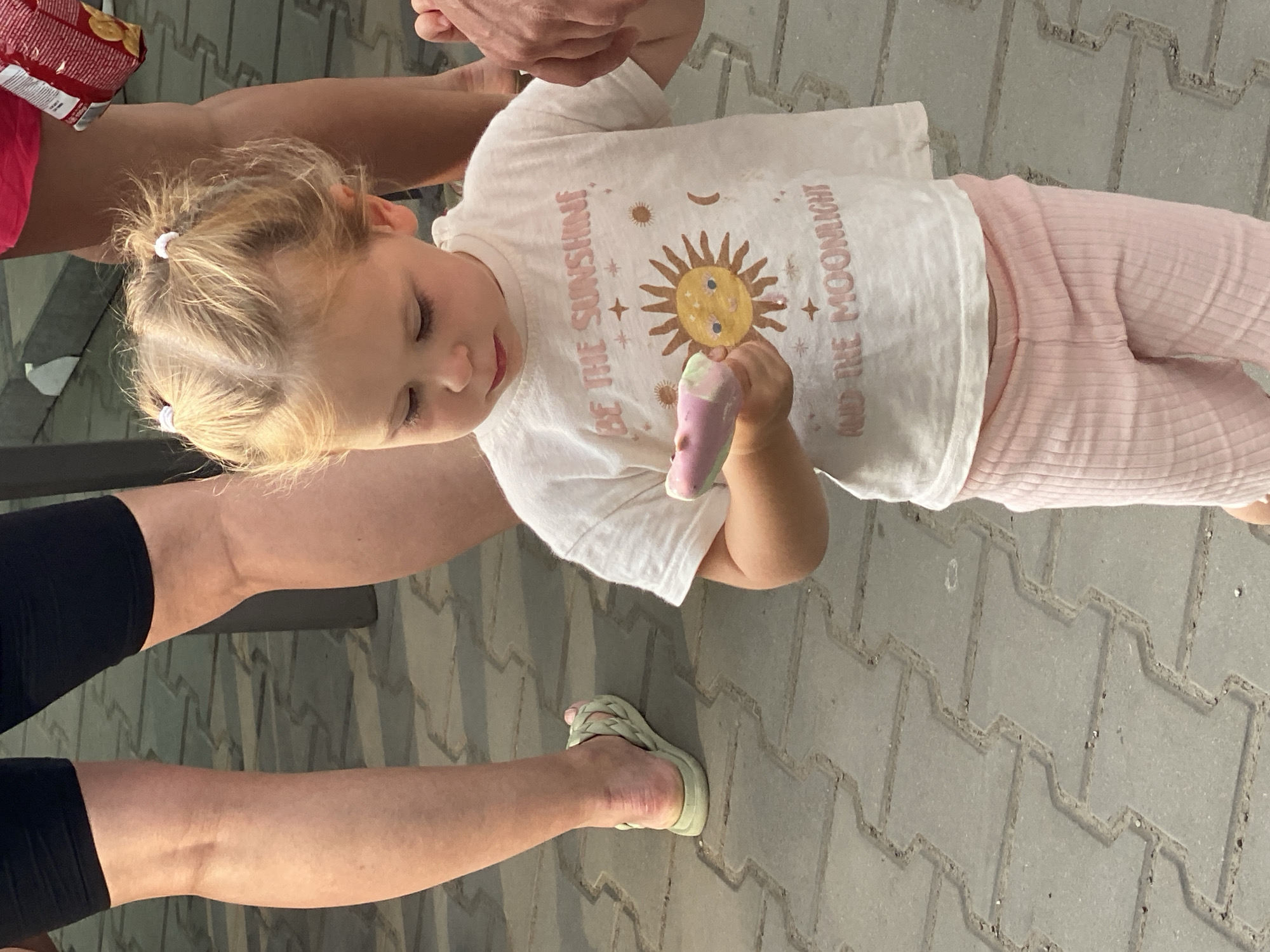 In Terebleche, Ukraine, a young blonde girl with her hair in pigtails is standing on a paved pathway. She is wearing a white t-shirt with a sun and moon graphic, and pink ribbed leggings. In her left hand, she holds a pink and white popsicle. The girl is looking down at the popsicle. Her legs are visible, wearing black shorts and light green sandals. In the background, other people's legs are visible, one pair wearing black shorts and another wearing bright pink shorts. The pathway is made of interlocking grey pavers.
