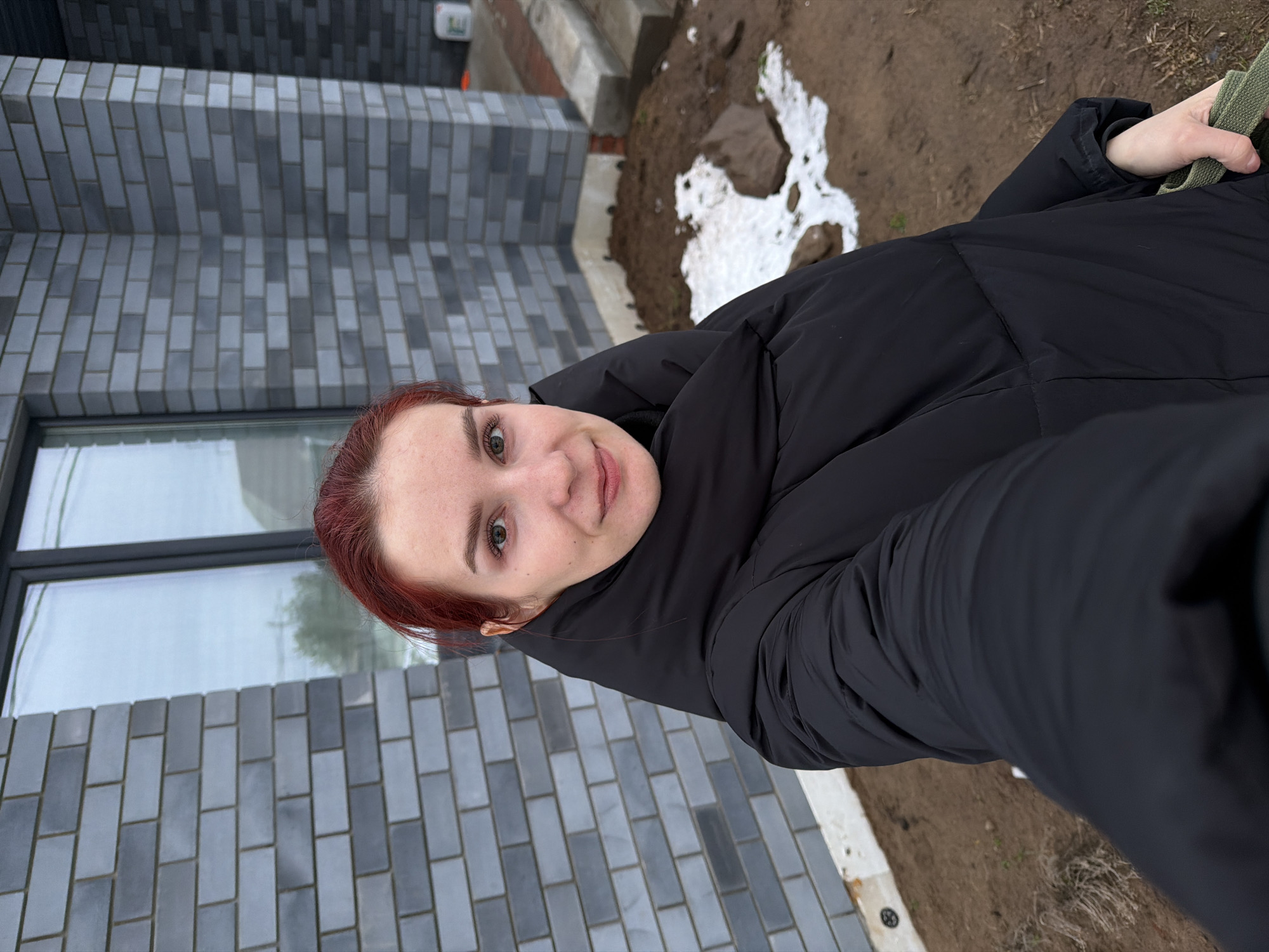 A person wearing a black puffy jacket stands outdoors in front of a building with a grey brick facade and a window. Patches of melting snow and dirt are visible on the ground. A strap of a bag is visible. The location is likely the city of Chaikovsky, Russia.