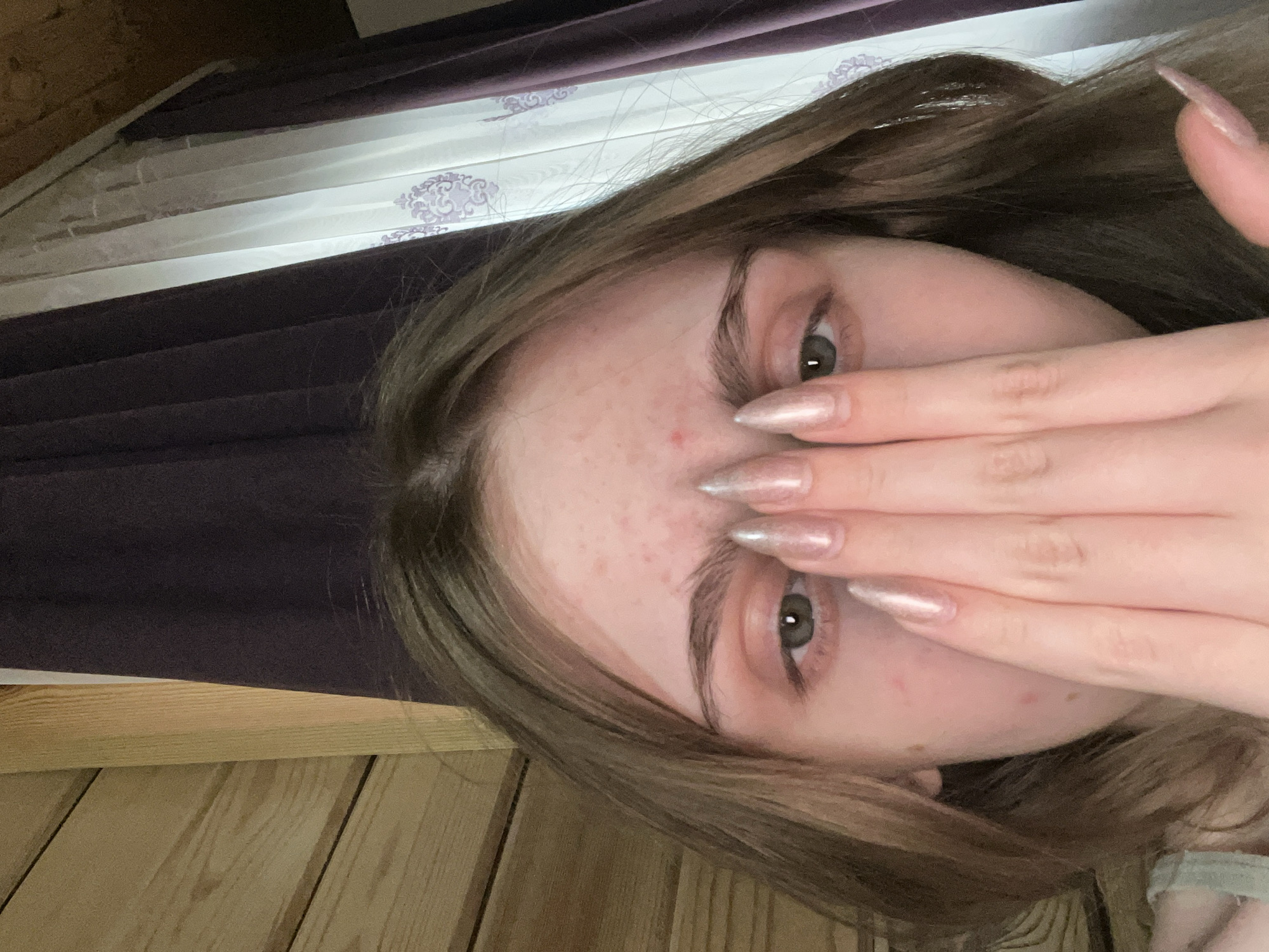 A close-up selfie shows a young person's face partially obscured by their hand. The person has fair skin with some blemishes and light brown hair. They are wearing long, metallic-pink fingernails. The background consists of wooden paneling and draped curtains, suggesting an indoor setting. There is no information in the image to indicate a location in Ashkadar, Russia.