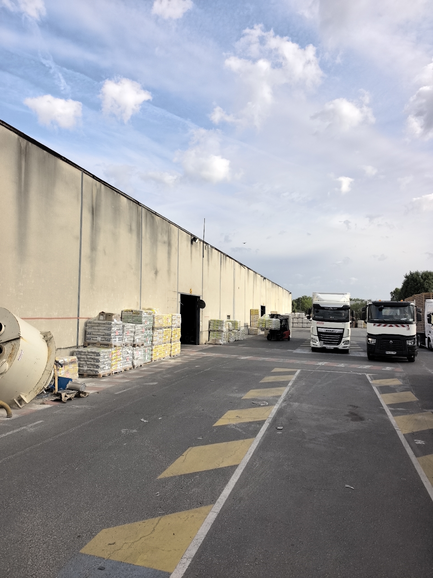 The image depicts a loading dock area outside a large, beige industrial building in Bonneuil-sur-Marne, France. The building has a dark roofline and a row of exterior lights. In front of the building, several pallets of bagged materials, likely cement or similar construction supplies, are stacked and wrapped in plastic. To the left, a large, yellow industrial mixer is positioned.
In the foreground and mid-ground, the asphalt surface is marked with yellow and white directional lines and crosswalk markings. Several trucks are present in the area. Two white semi-trailer trucks are parked, one with "DAF" branding and the other with a red and white striped marking on its cab. A red forklift is also visible near some stacked pallets. The sky is partly cloudy with a mix of blue sky and white clouds. Some trees are visible in the background to the right.