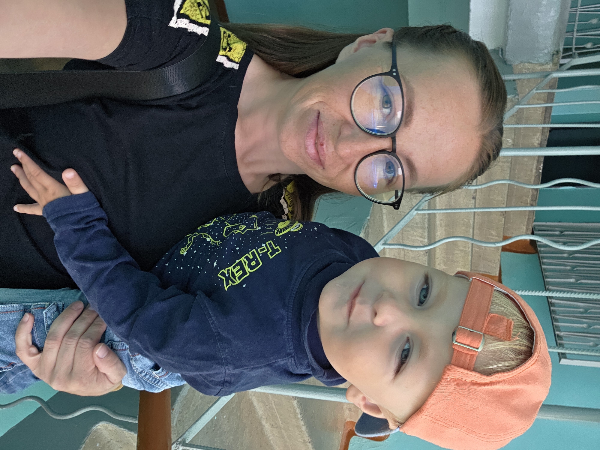 A woman with blonde hair pulled back and glasses is holding a young child with blonde hair and a peach-colored baseball cap. The woman is wearing a black t-shirt with yellow and white accents on the shoulder. The child is wearing a dark blue long-sleeved shirt with a "T-REX" graphic and denim jeans. They are standing in front of a staircase with teal-colored walls and a metal railing. The location is identified as the stairwell of an apartment building in Chaikovsky, Russia. The lighting suggests it is daytime.
