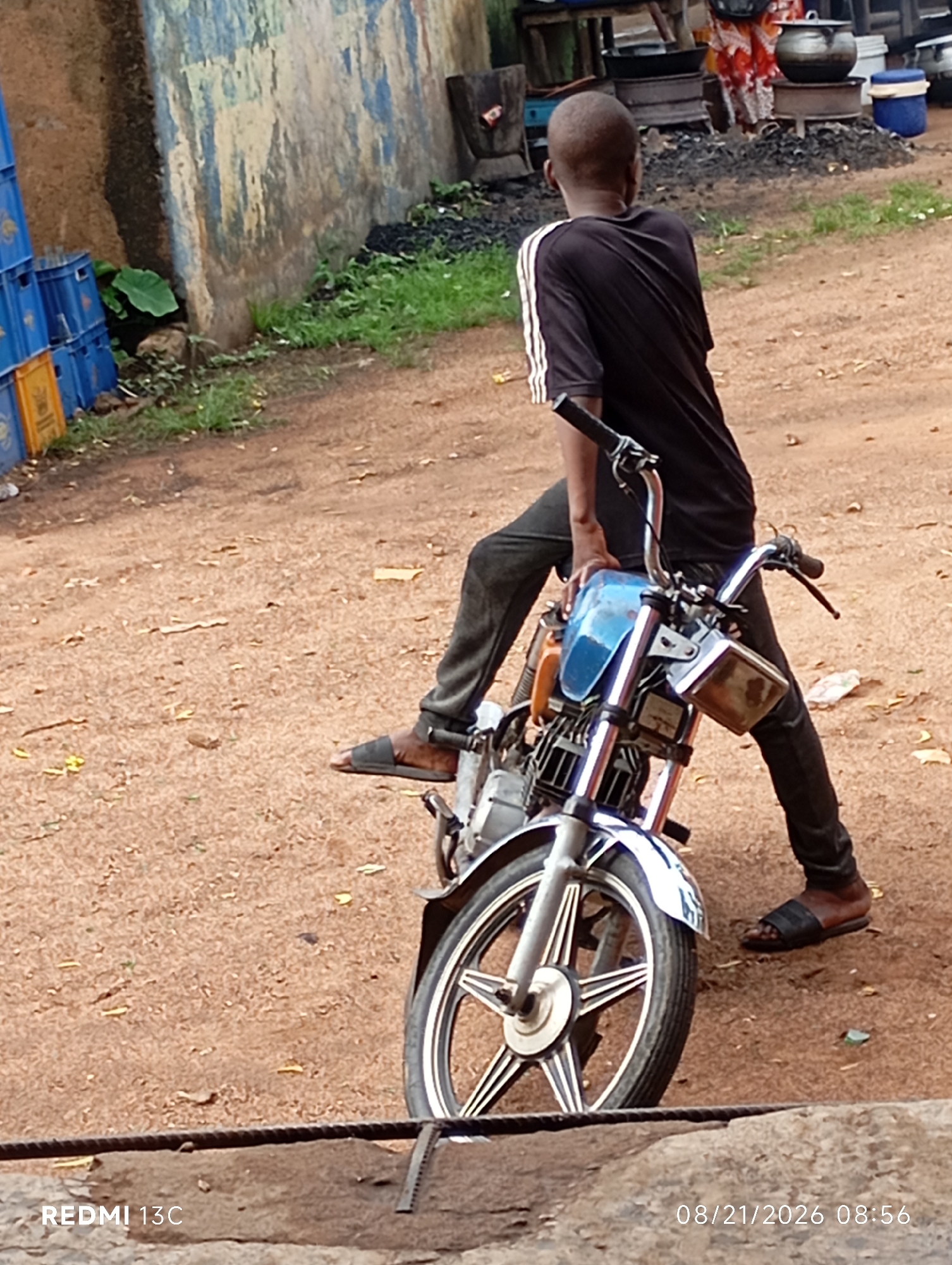 A young person wearing a black t-shirt with white stripes on the sleeves and black pants sits on a blue and silver motorcycle in an outdoor environment. The motorcycle is the primary focus of the image. The ground is covered in reddish-brown dirt and scattered dry leaves. In the background, there's a weathered, multi-colored wall with blue and white paint, and stacks of blue plastic crates. To the right, there are outdoor cooking setups with pots and charcoal. The image is dated "08/21/2026 08:56" and tagged "REDMI 13C."