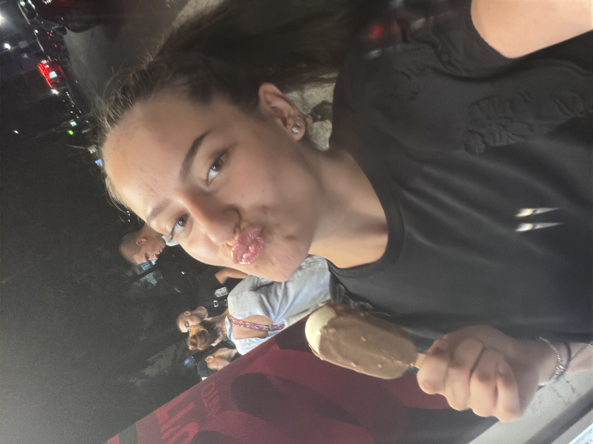 The image, taken at night in Burgas, Bulgaria, shows a young woman holding an ice cream cone and making a kissing face. She is wearing a black t-shirt. In the background, other people are visible, and there is a street scene with cars and lights.