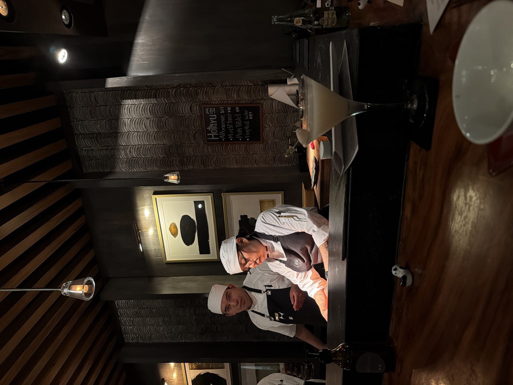Here is a concise description of the image:
The image shows the interior of a restaurant. There are two chefs working at a counter. A cocktail glass is near the edge of the counter, and a bowl is on a wooden table. There are framed pictures on the wall, and a menu. The location appears to be "Hotaru" based on the menu. There are bottles of Topo Chico sparkling mineral water on the counter. There are interesting architectural features of the restaurant including the wall tiling.