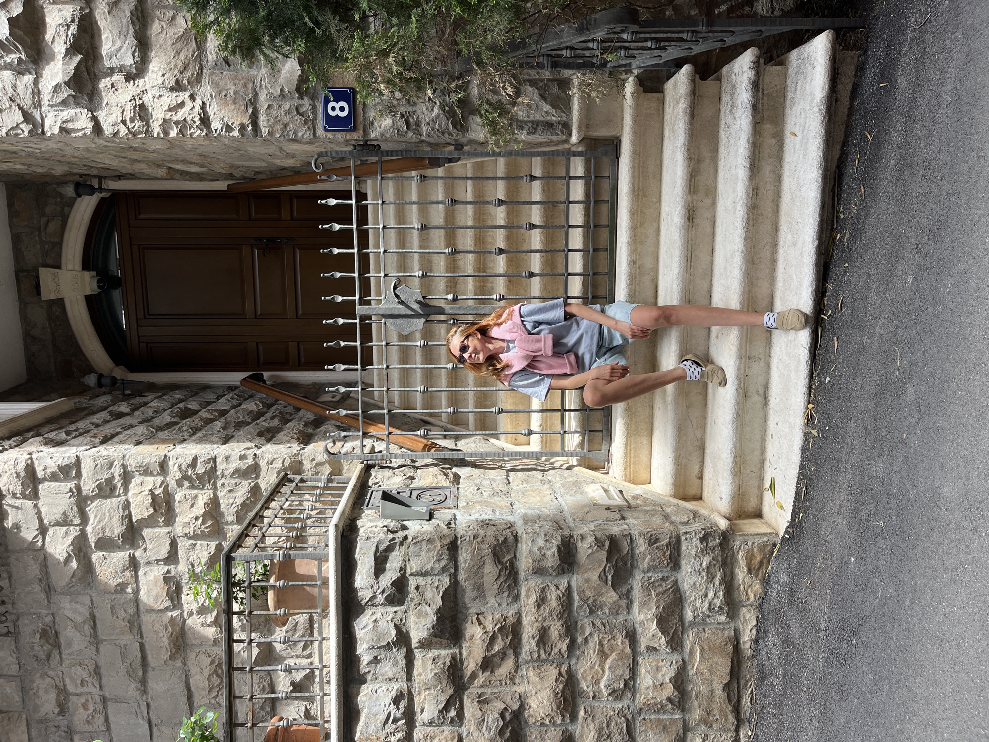 A woman sits on the white stone steps in front of a building constructed of rough-cut stone blocks. The woman wears sunglasses, a light blue t-shirt, light denim shorts, and tan sneakers with a white and black checkered pattern at the heel. A pink sweater is draped around her neck. The entrance is protected by a dark iron gate, and a small blue number "8" is affixed to the wall above it. To the left of the gate, a smaller wrought iron railing borders a section of stonework with a clay pot and small plants. The stairs run parallel to a narrow paved road. Based on the architecture and urban details, the location is likely in the city of Budva, Montenegro.