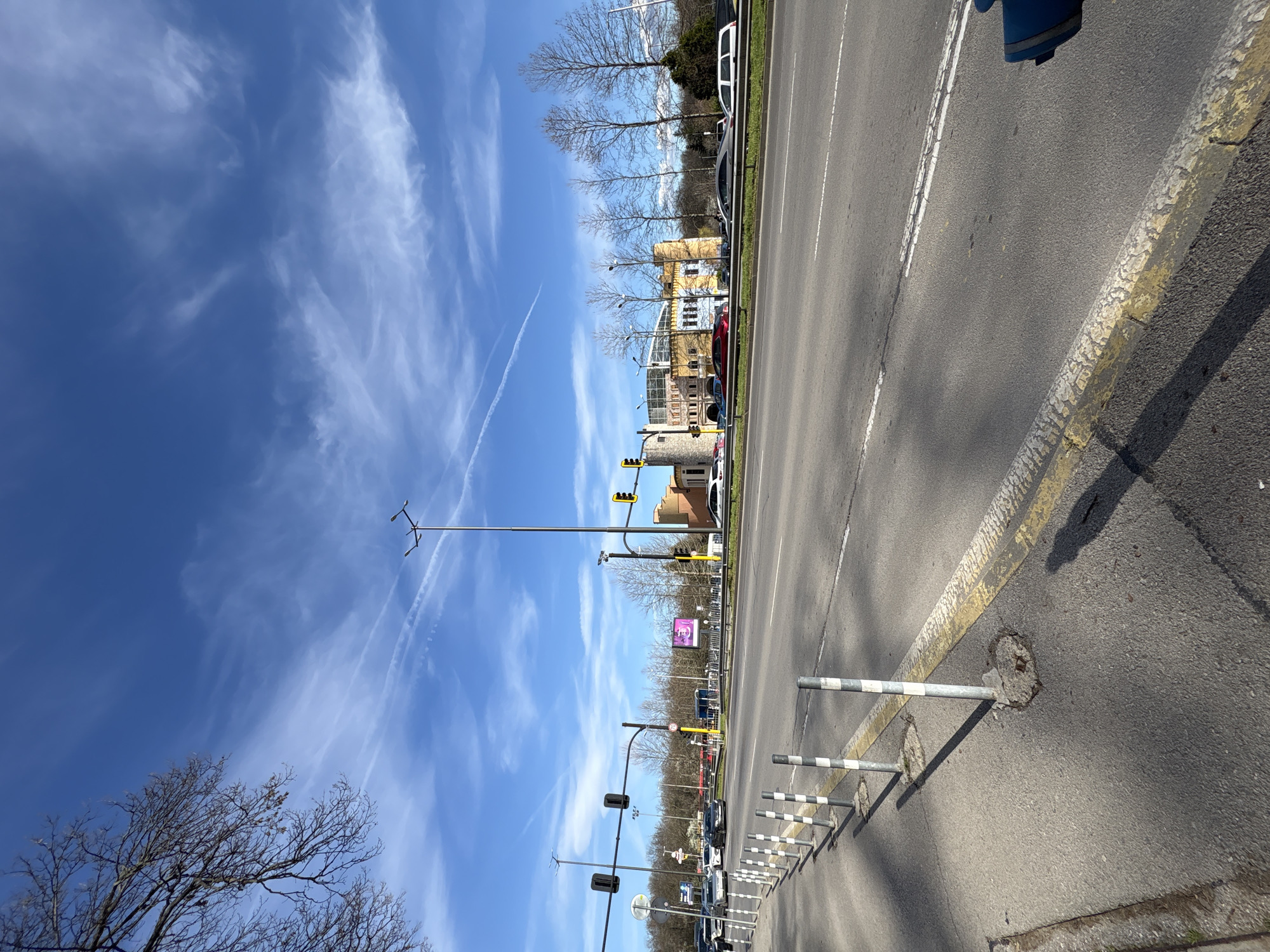 The image shows a street scene in Sofia, Bulgaria. The view is oriented towards the sky and clouds. In the foreground, there is a portion of a street with a yellow-painted curb and a series of metal barriers with white stripes. A grey asphalt road extends into the distance. Along the roadside, there are trees, traffic lights, and buildings. In the background, there are several buildings of various heights and colors. The sky is blue with streaks of white clouds.