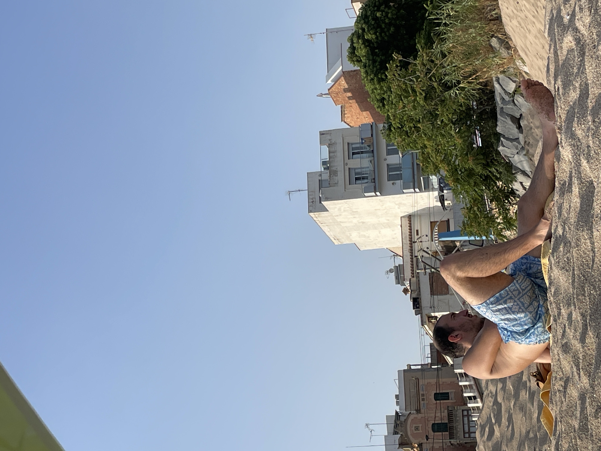 A man is resting on a sandy beach in Canet de Mar, Spain. He is lying on his back, propped up on his forearms, and looking to his right. He is wearing blue patterned swim trunks. The beach sand is light brown and textured. Behind him, there are several buildings, some of which are multi-story and appear to be residential or hotels. There is also dense green foliage visible behind the buildings, and a clear blue sky above. A yellow towel is partially visible beneath the man.