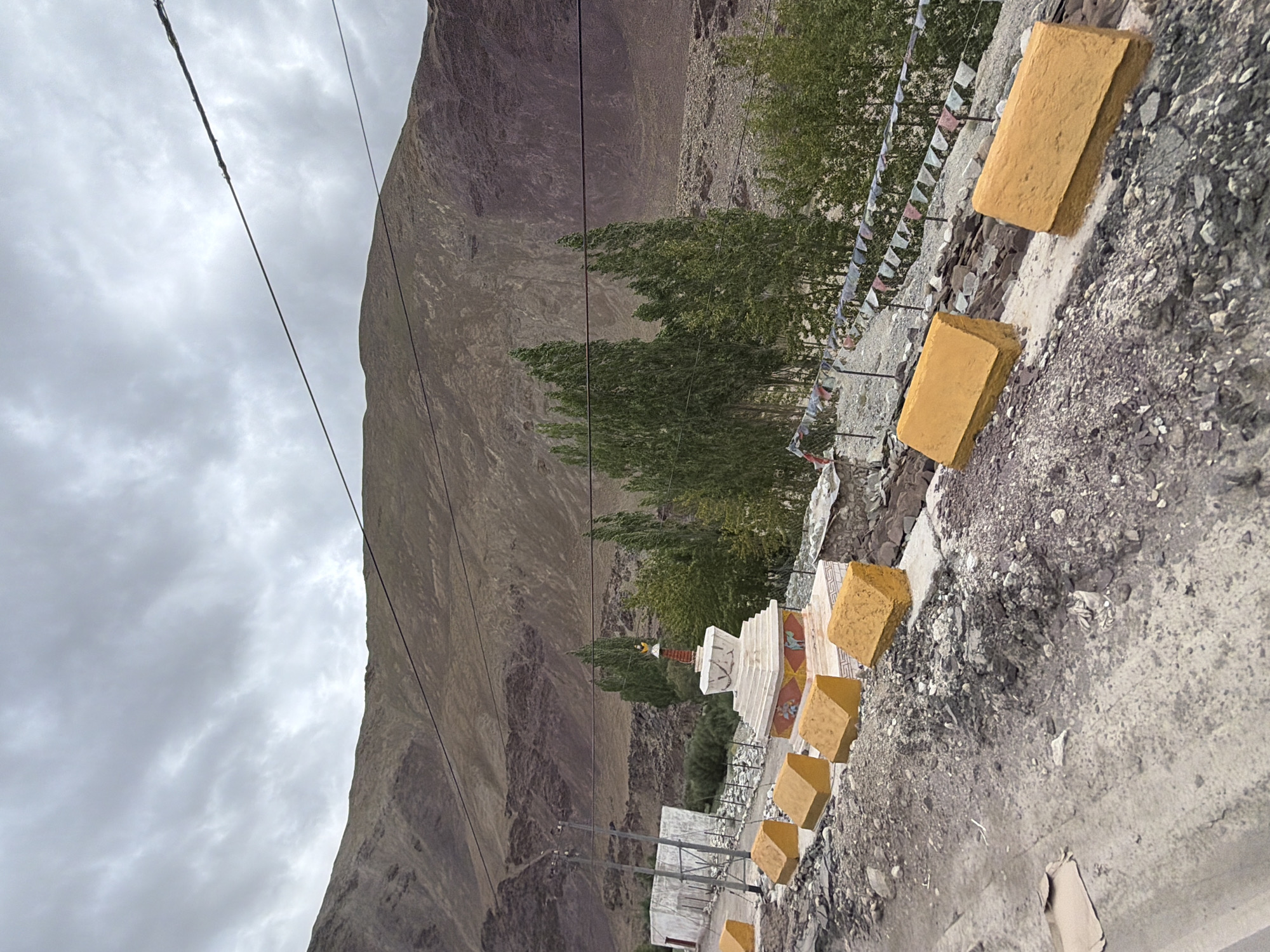The image depicts a roadside scene in Kiari, India, under a cloudy sky. In the foreground, a dirt road is visible, with several rectangular, yellow barriers lining its edge. Beyond the road, a white stupa with red and white decorative bands sits prominently, surrounded by a low stone wall. A line of tall, slender green trees stands behind the stupa, with more vegetation and rocky terrain stretching towards a large, barren mountain in the background. Power lines are strung across the sky. A white building with a red trim is partially visible to the left of the stupa, with a metal fence and power poles nearby. Small prayer flags are strung on a fence to the right of the stupa.